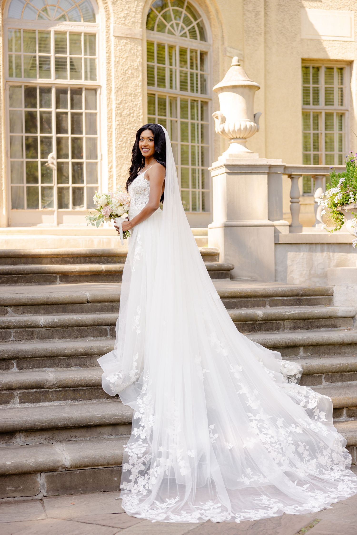 A dramatic cathedral-length wedding veil trails down stone steps at an elegant historic venue.