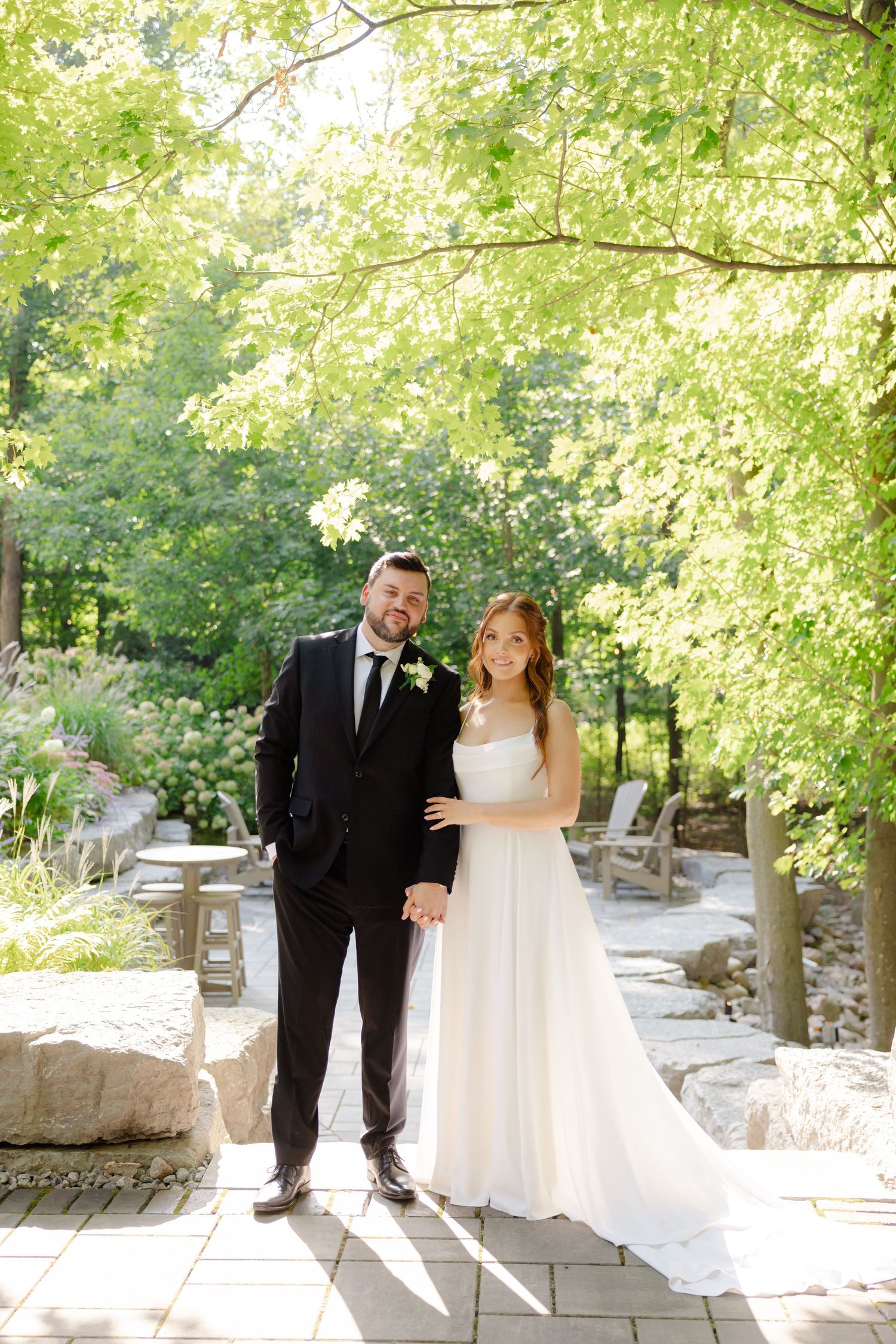 A full-length wedding portrait on a stone pathway under green trees with dappled sunlight.