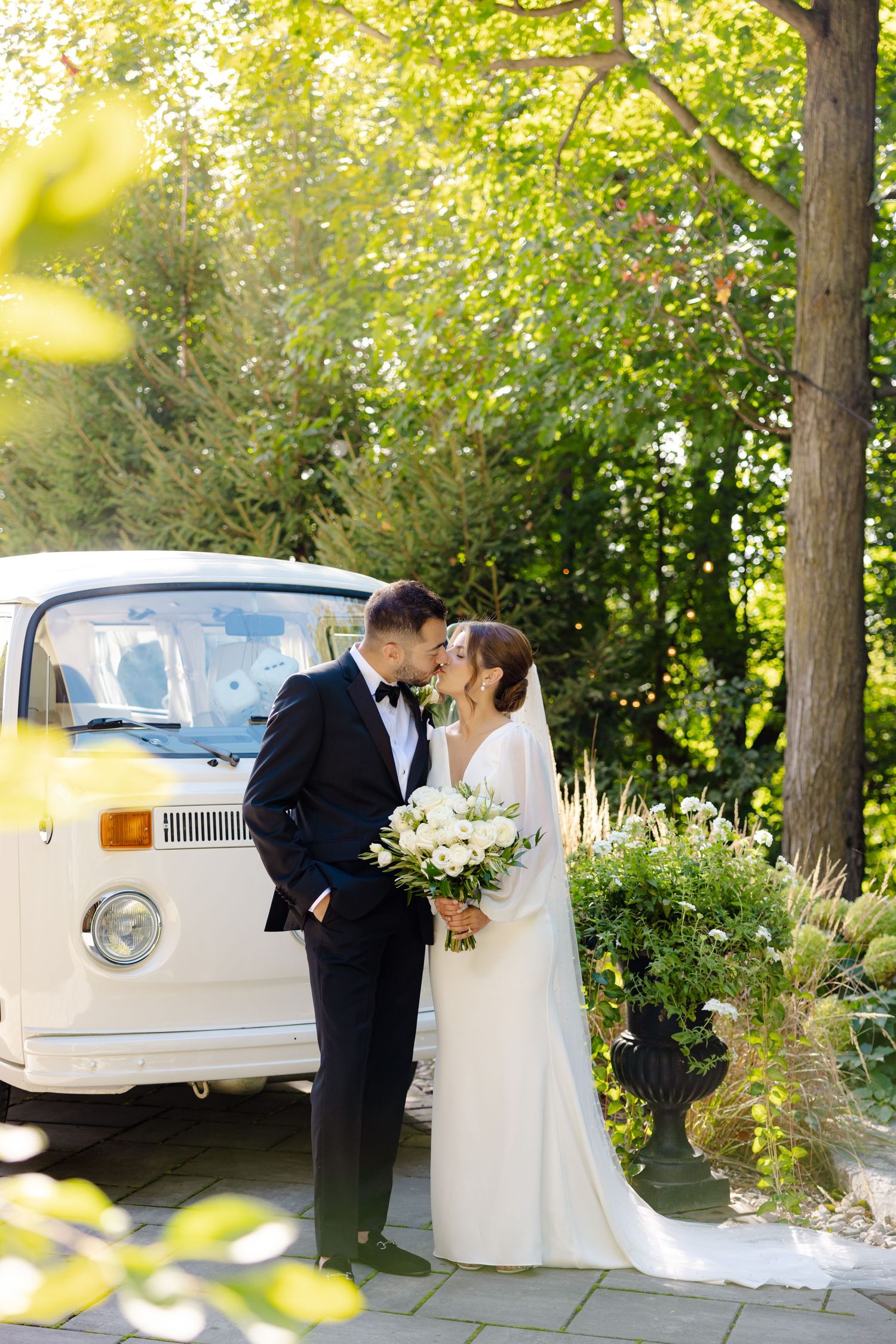 Intimate moment between newlyweds beside classic white VW van in a garden setting.