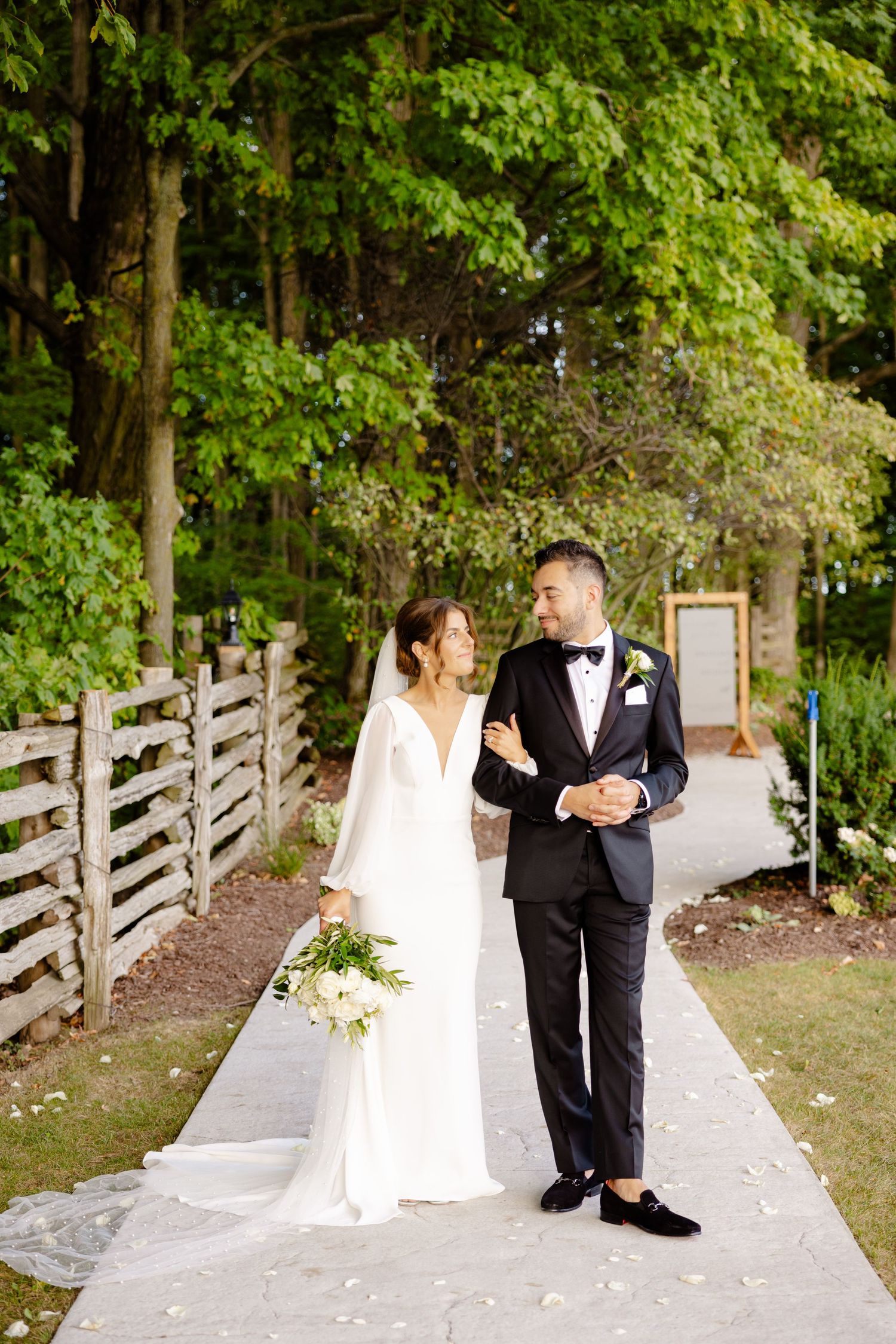 A wedding couple strolls along a scenic pathway lined with wooden fencing and trees.