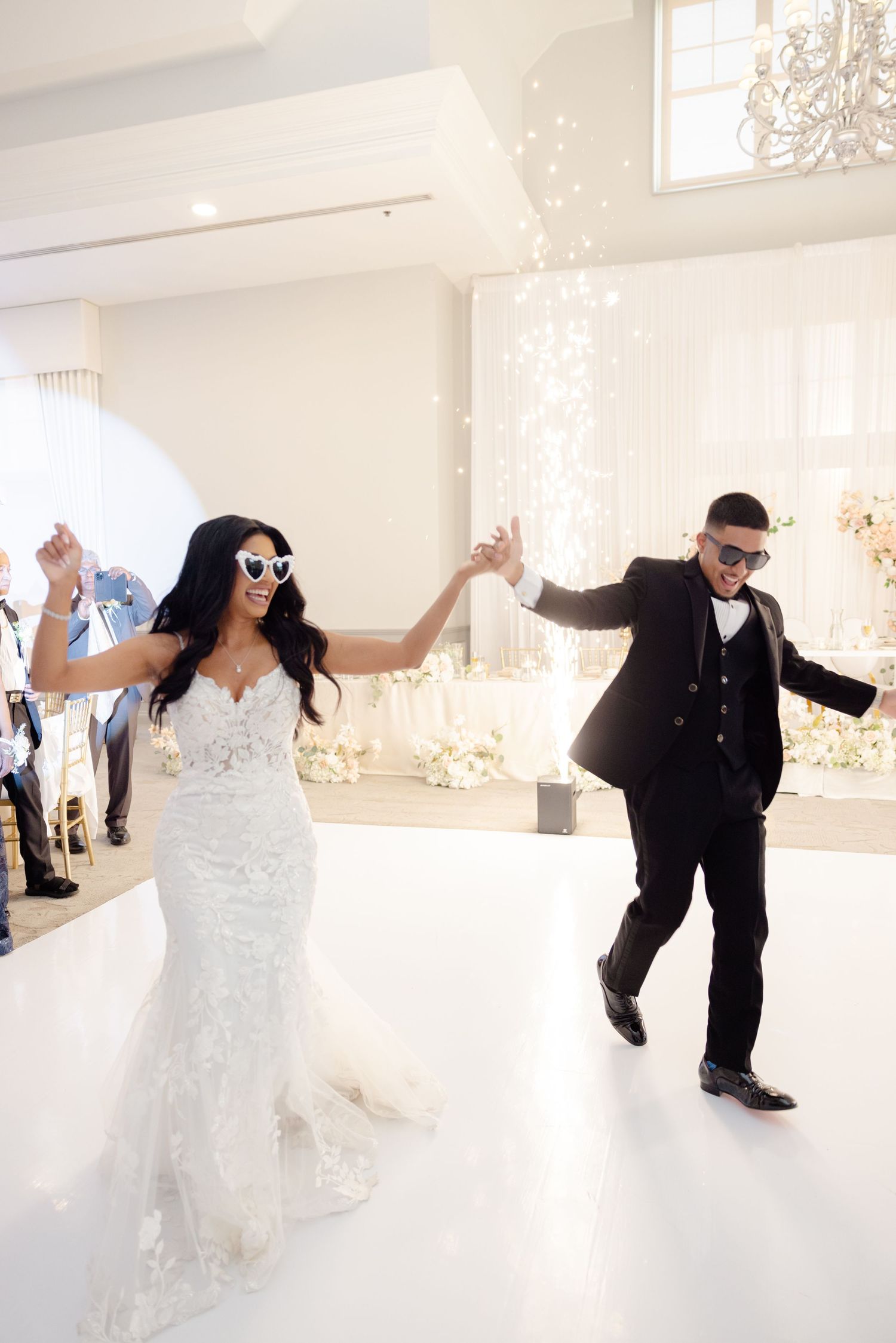 Energetic dance moves on a white platform during a wedding celebration with sparkly backdrop.