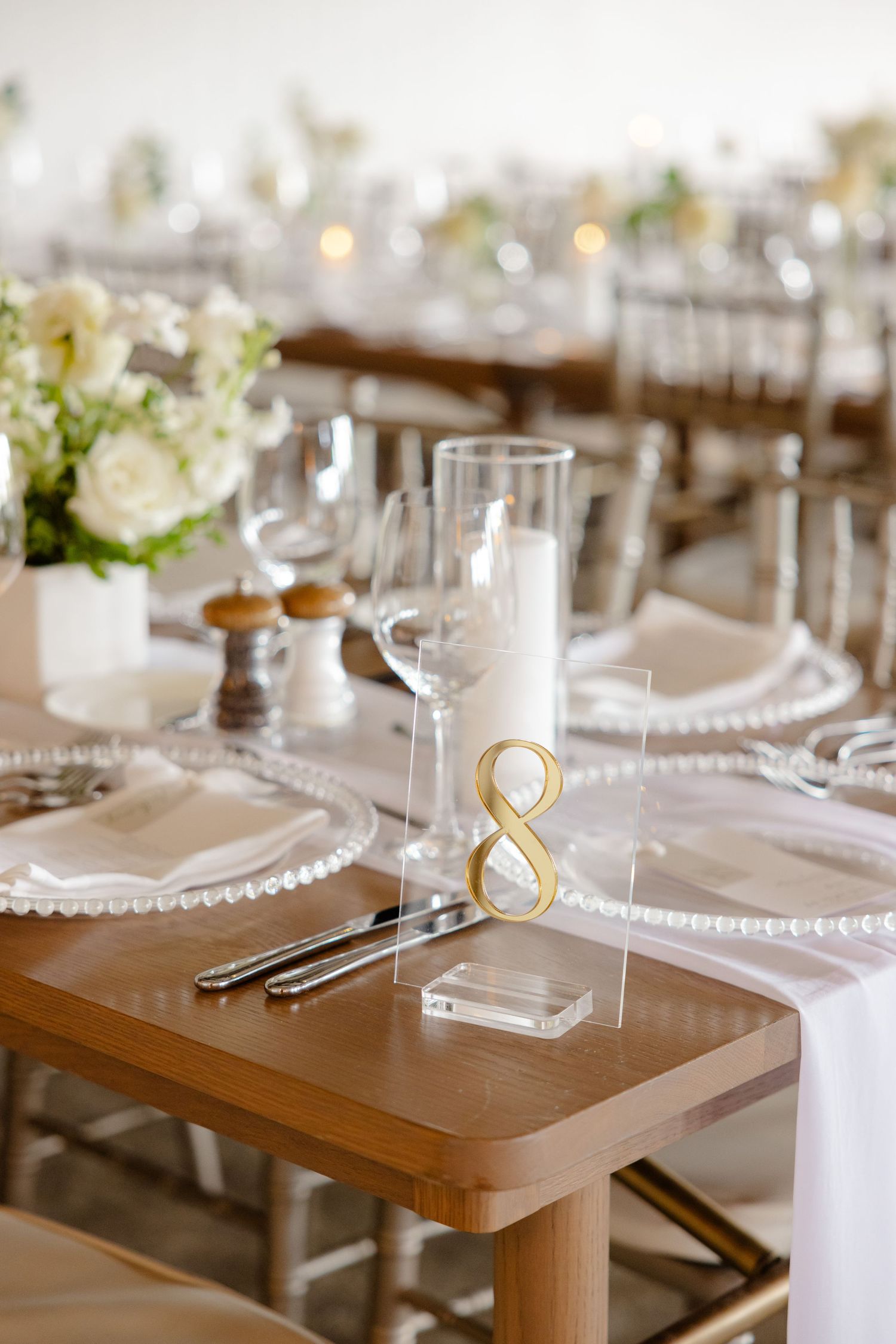 Table setting with gold number 8 stand, beaded charger plates, and white floral centerpiece.