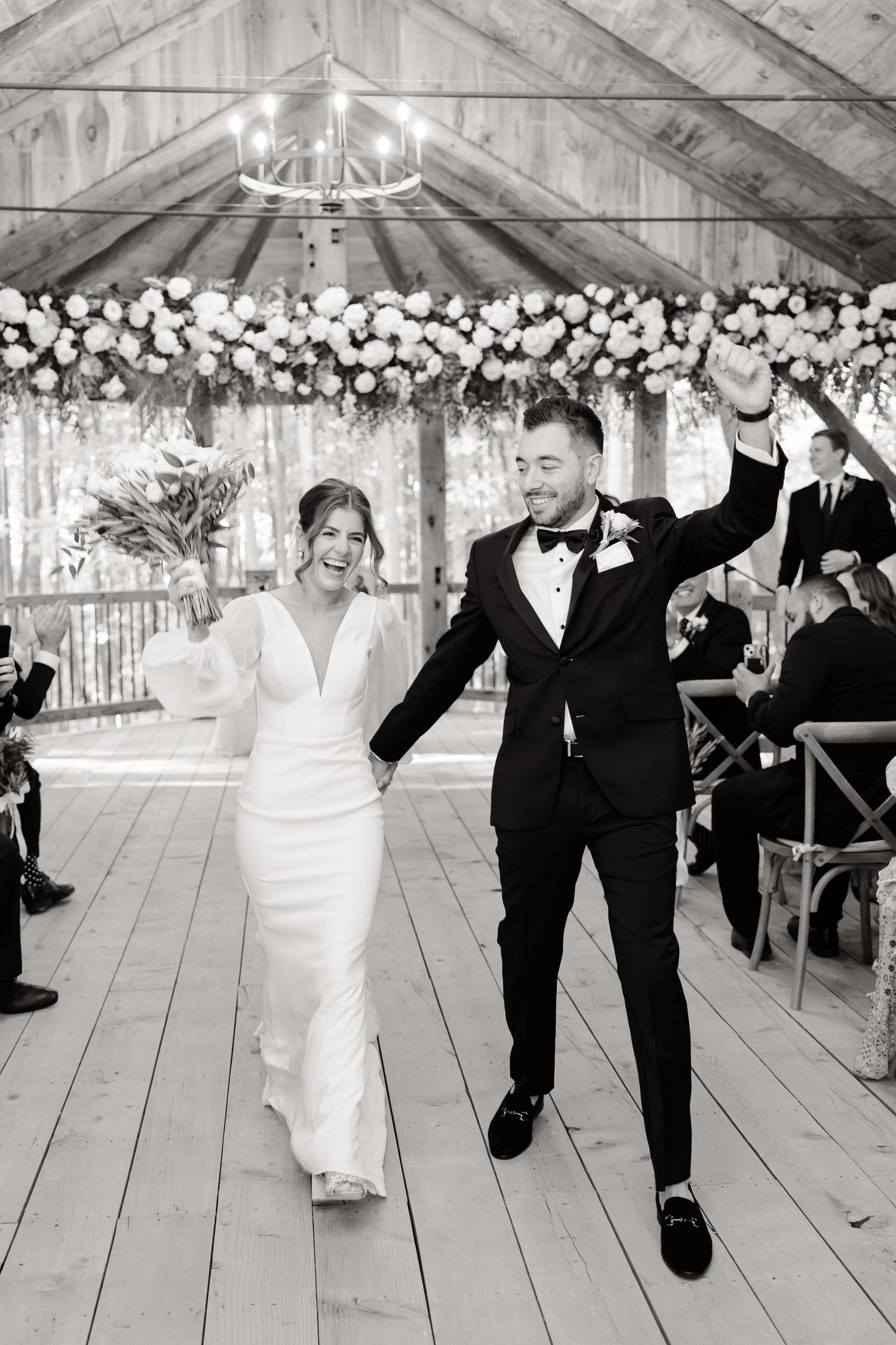 A joyful wedding recessional in black and white under a rustic wood pavilion with floral garlands.