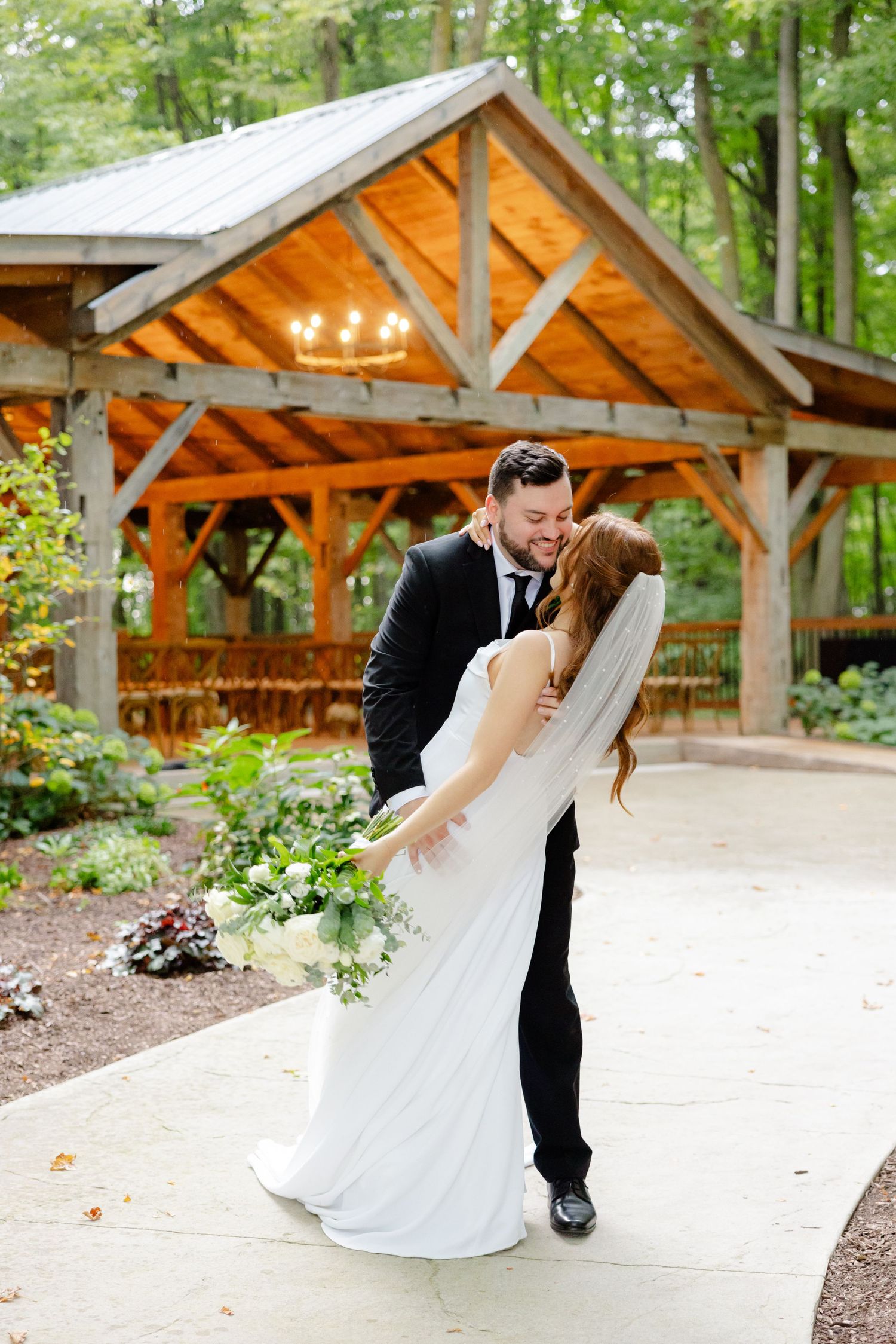 Newlyweds share romantic moment in front of rustic wooden pavilion with glowing lights in forest setting.