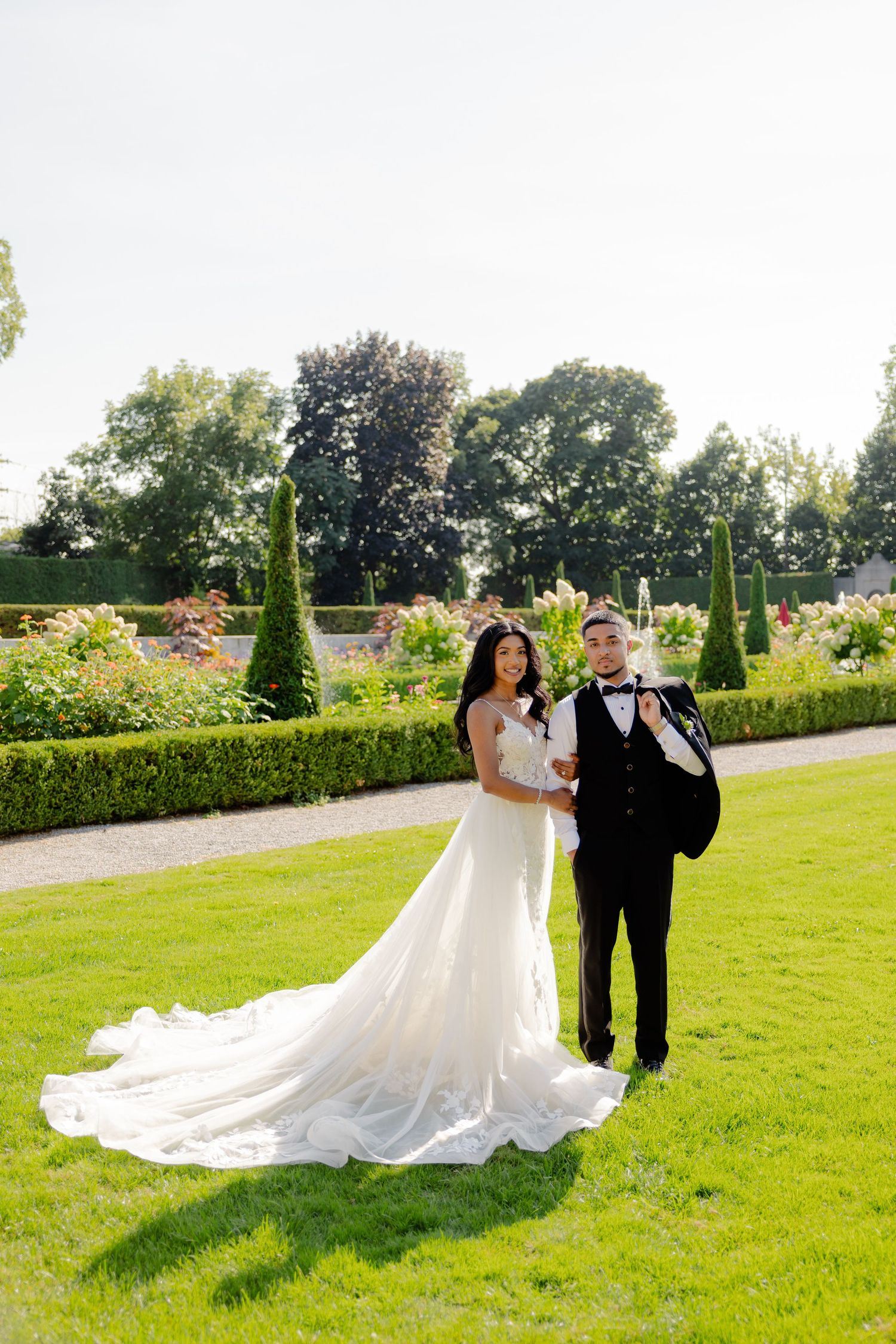 Elegant wedding portrait in a formal garden setting with manicured hedges and flowering plants.