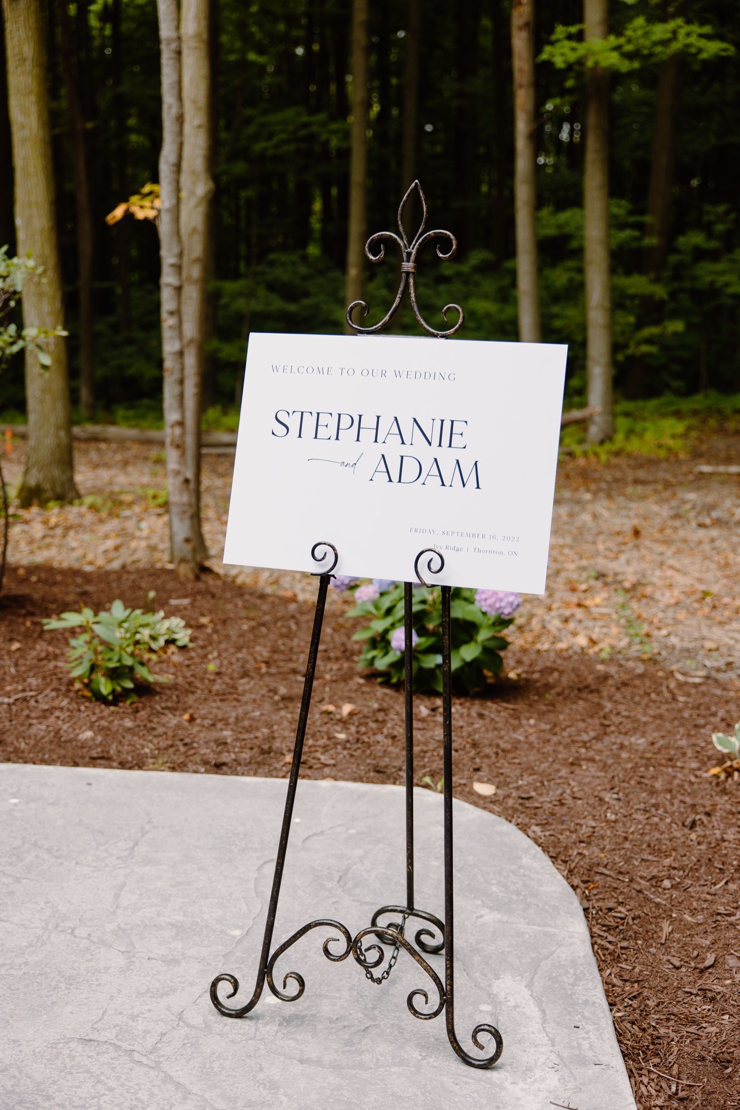 Ornate metal stand displays wedding signage against wooded forest background.