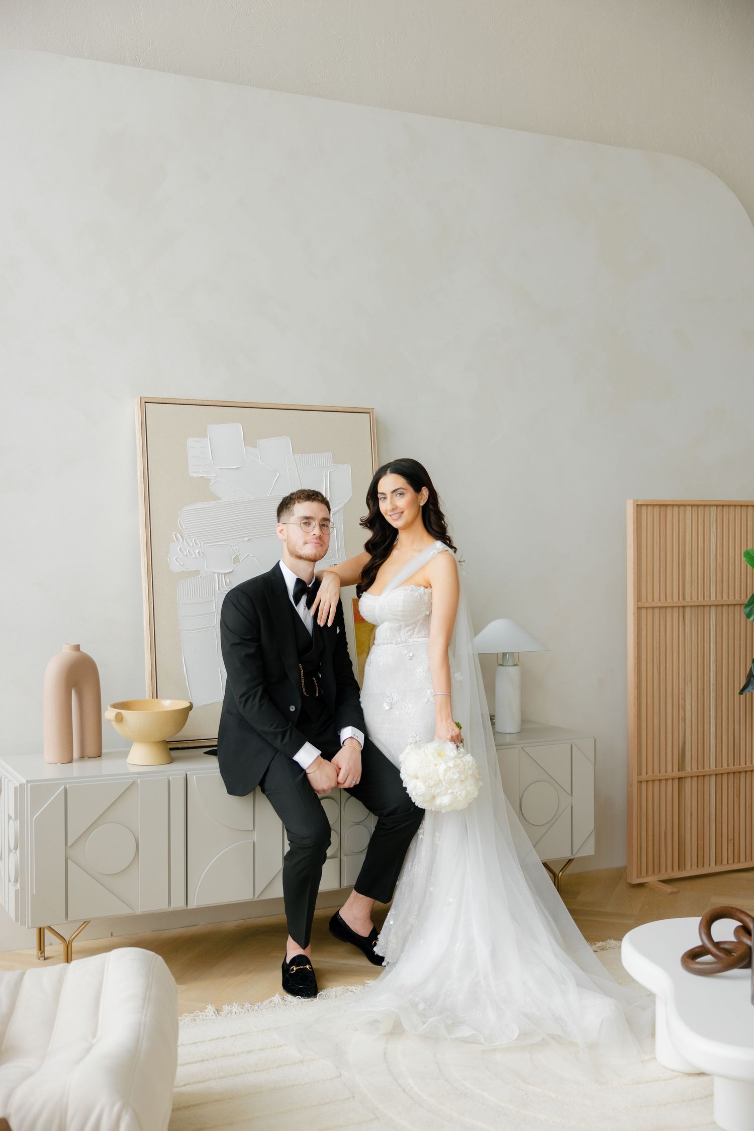 Stylish wedding couple poses casually against modern decor in bright white interior space.