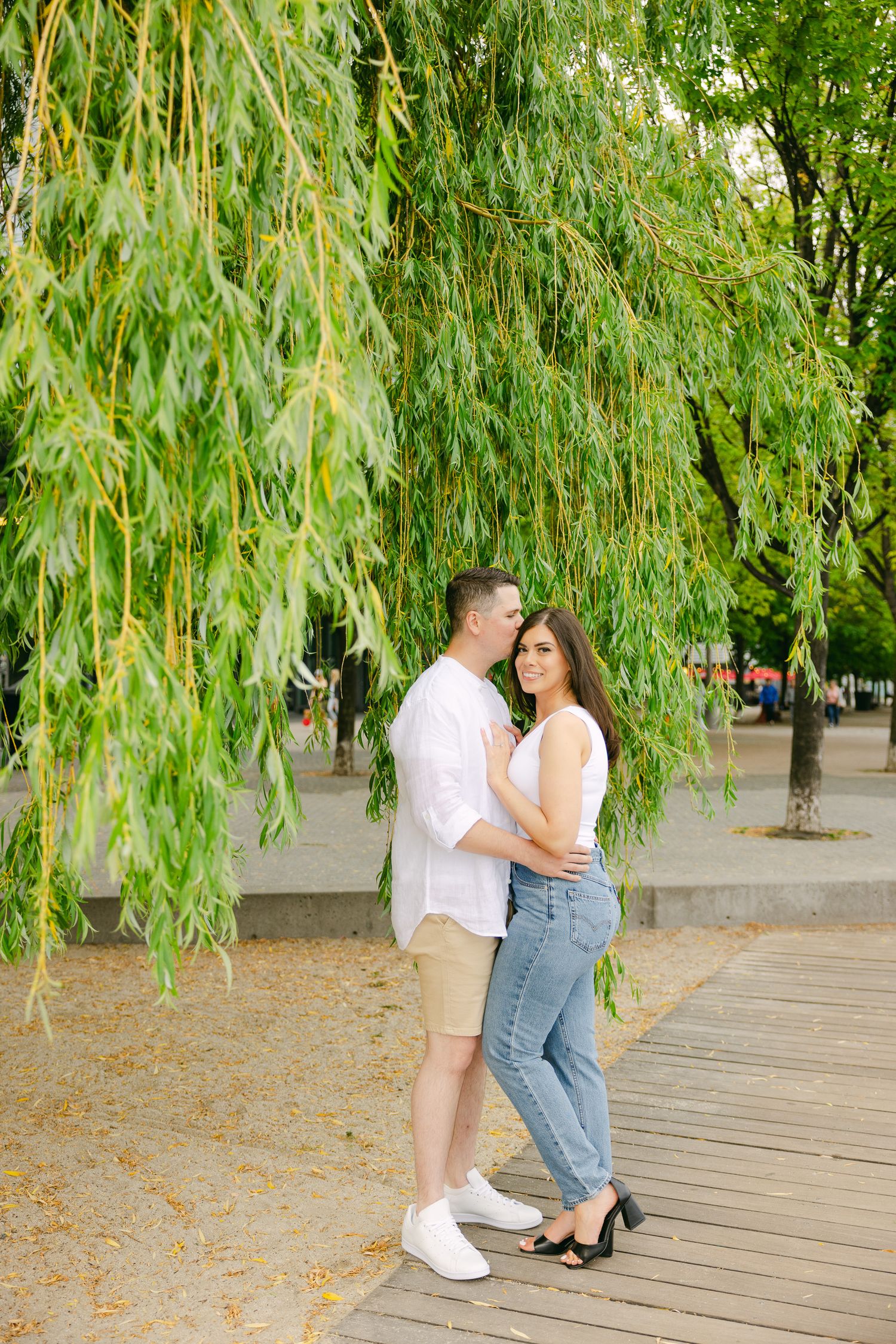 A couple sharing an intimate moment beneath hanging willow trees on a wooden boardwalk.