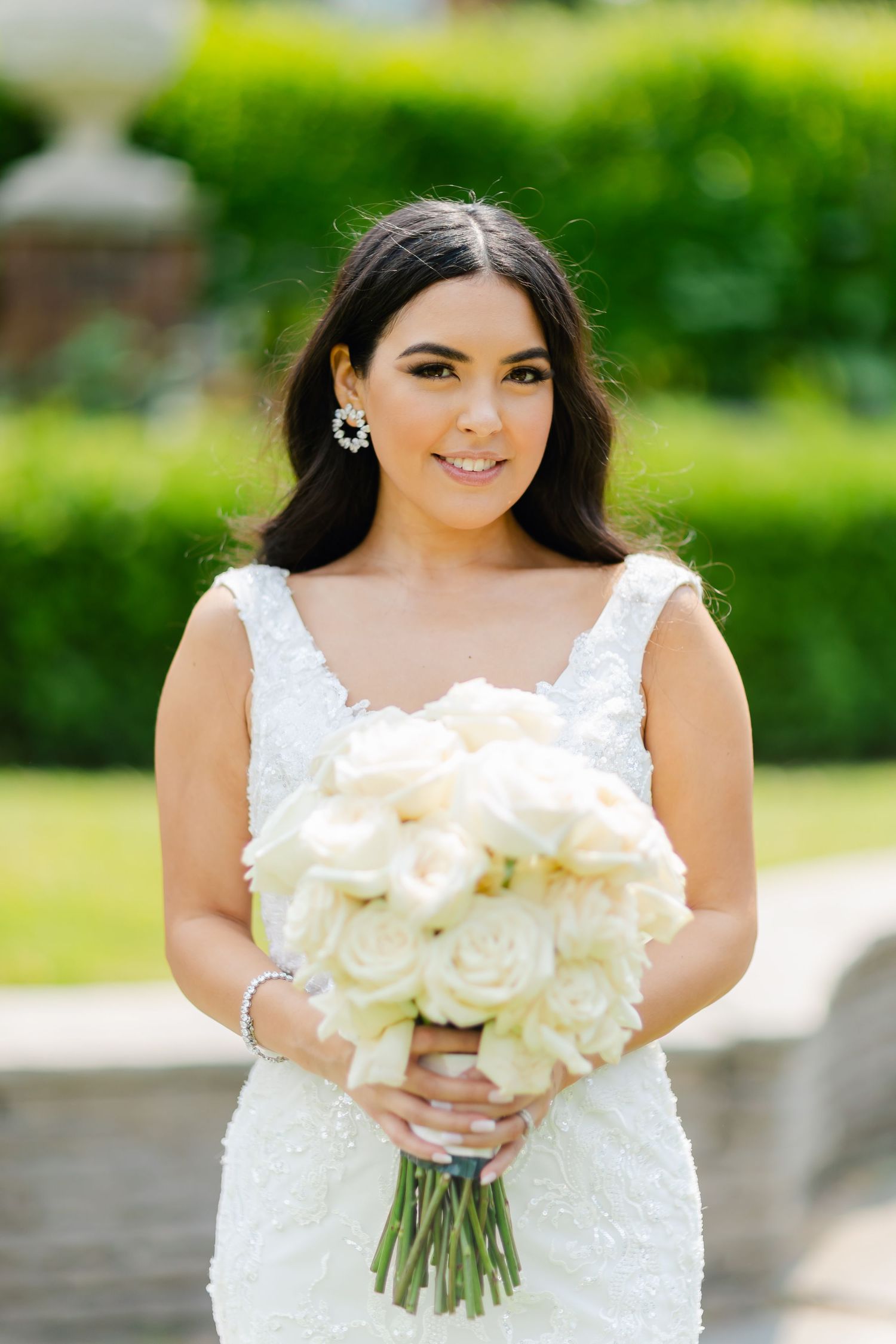 Bride holding a luxurious white rose bouquet poses in a lace wedding gown against a green garden backdrop.