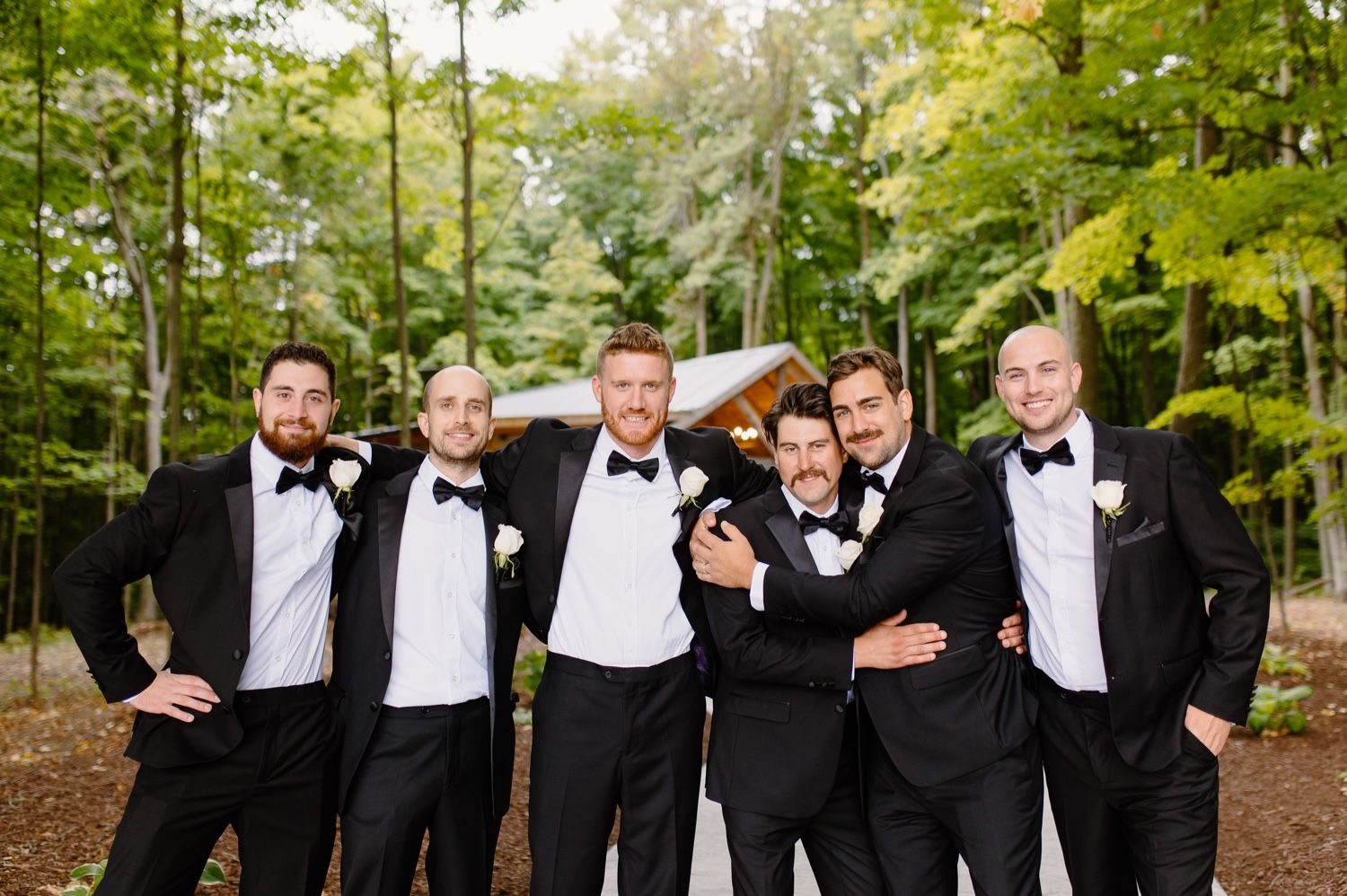 Group of groomsmen in black tuxedos posing casually and laughing together in a wooded setting.