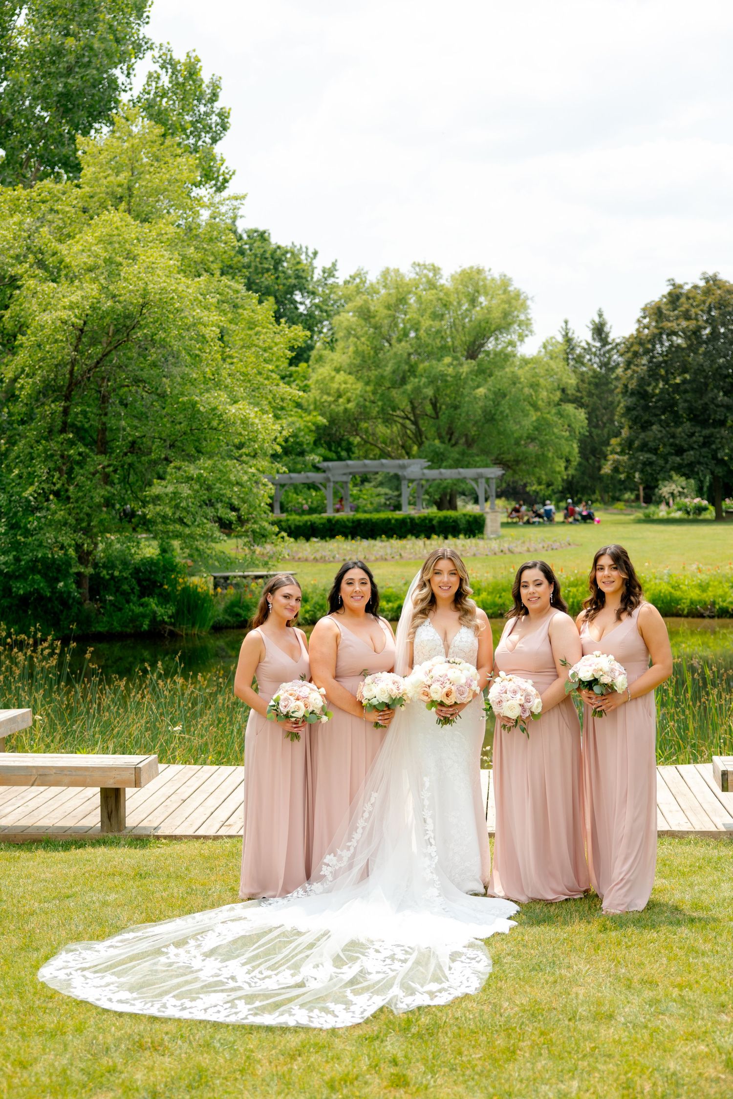 Bridal party in blush pink dresses poses on wooden boardwalk with dramatic cathedral veil in garden setting.