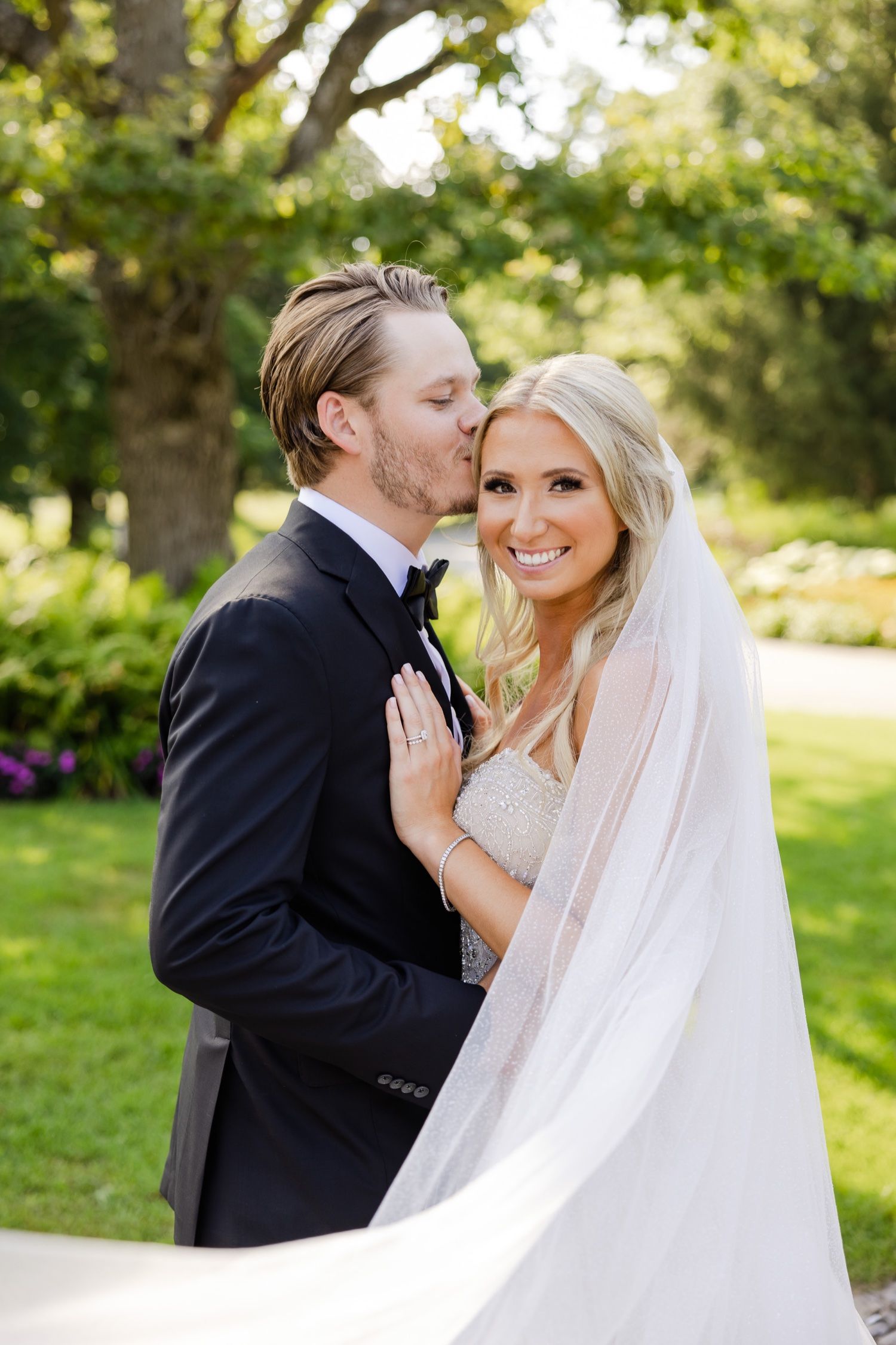An affectionate wedding moment captured in a garden with a flowing white veil and natural greenery.