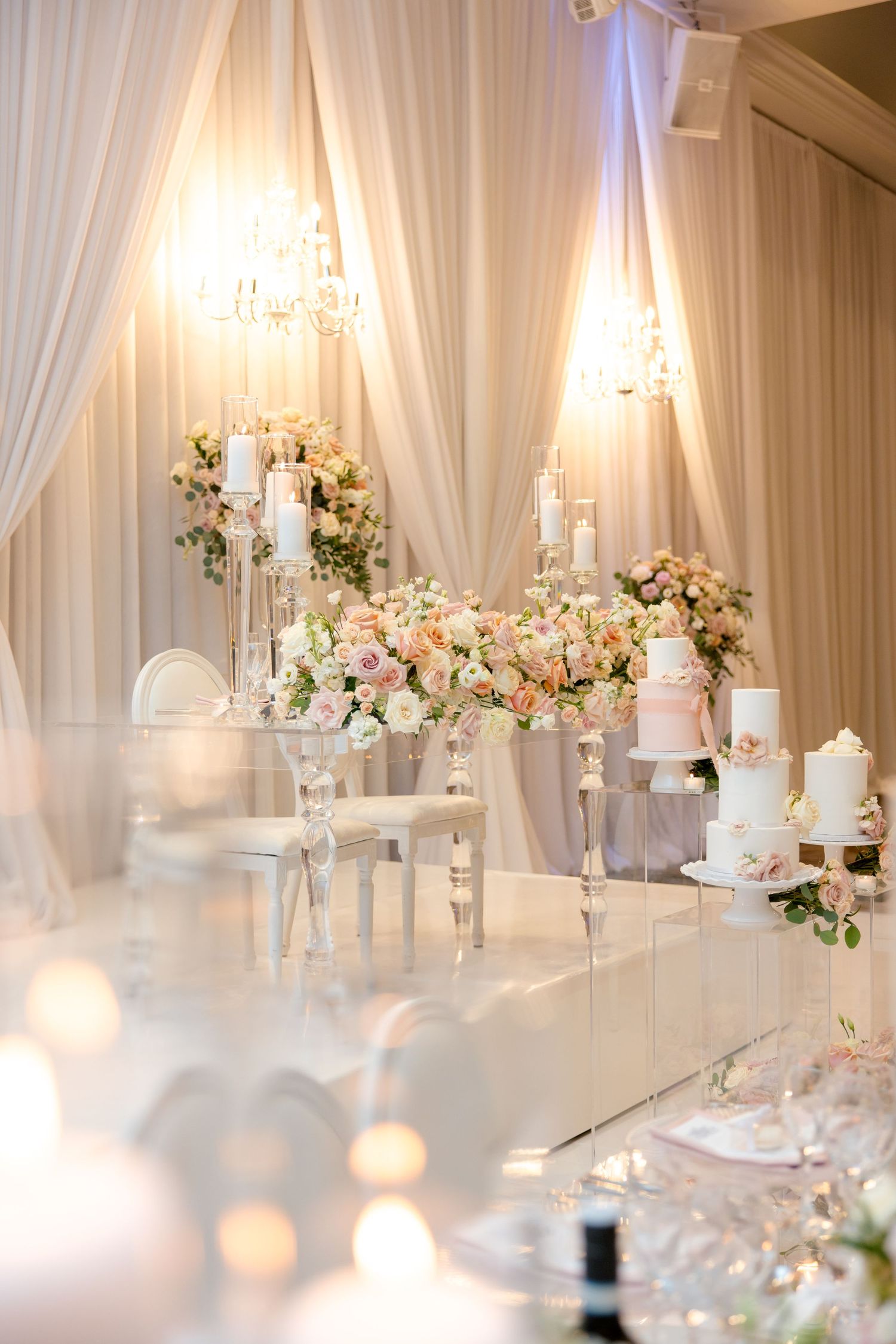 Luxurious white and blush wedding reception setup with crystal chandeliers, candles and floral centerpieces.