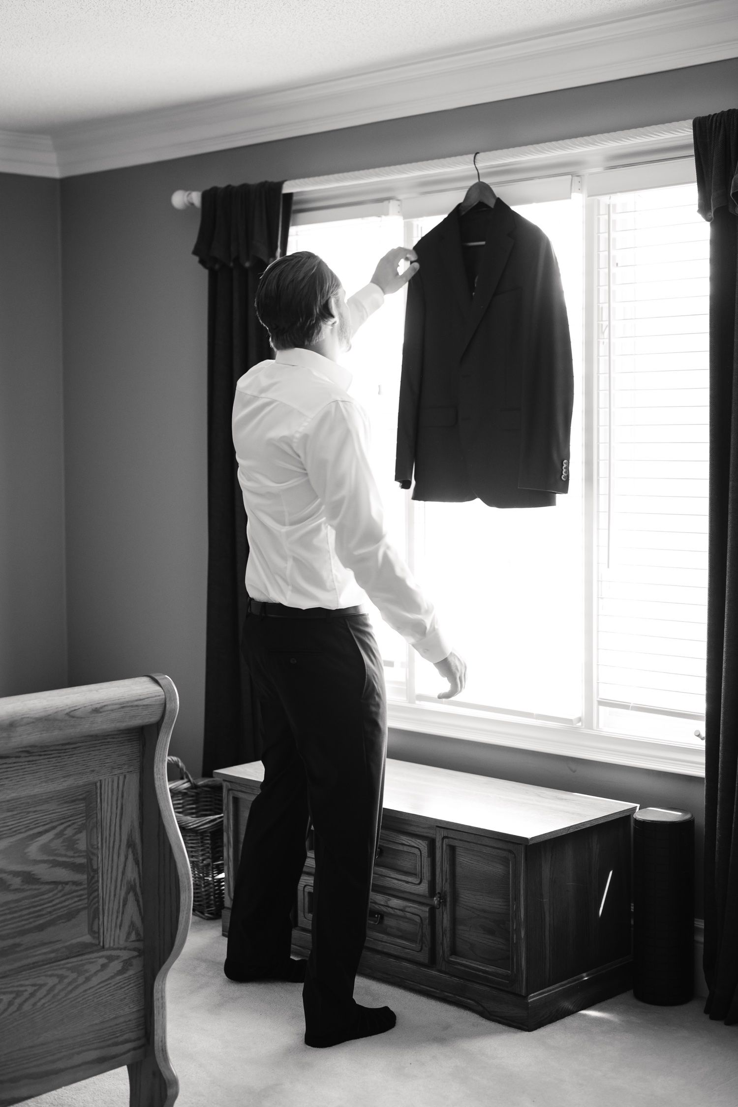 A person in white shirt reaches up to adjust a black suit jacket hanging by a window.
