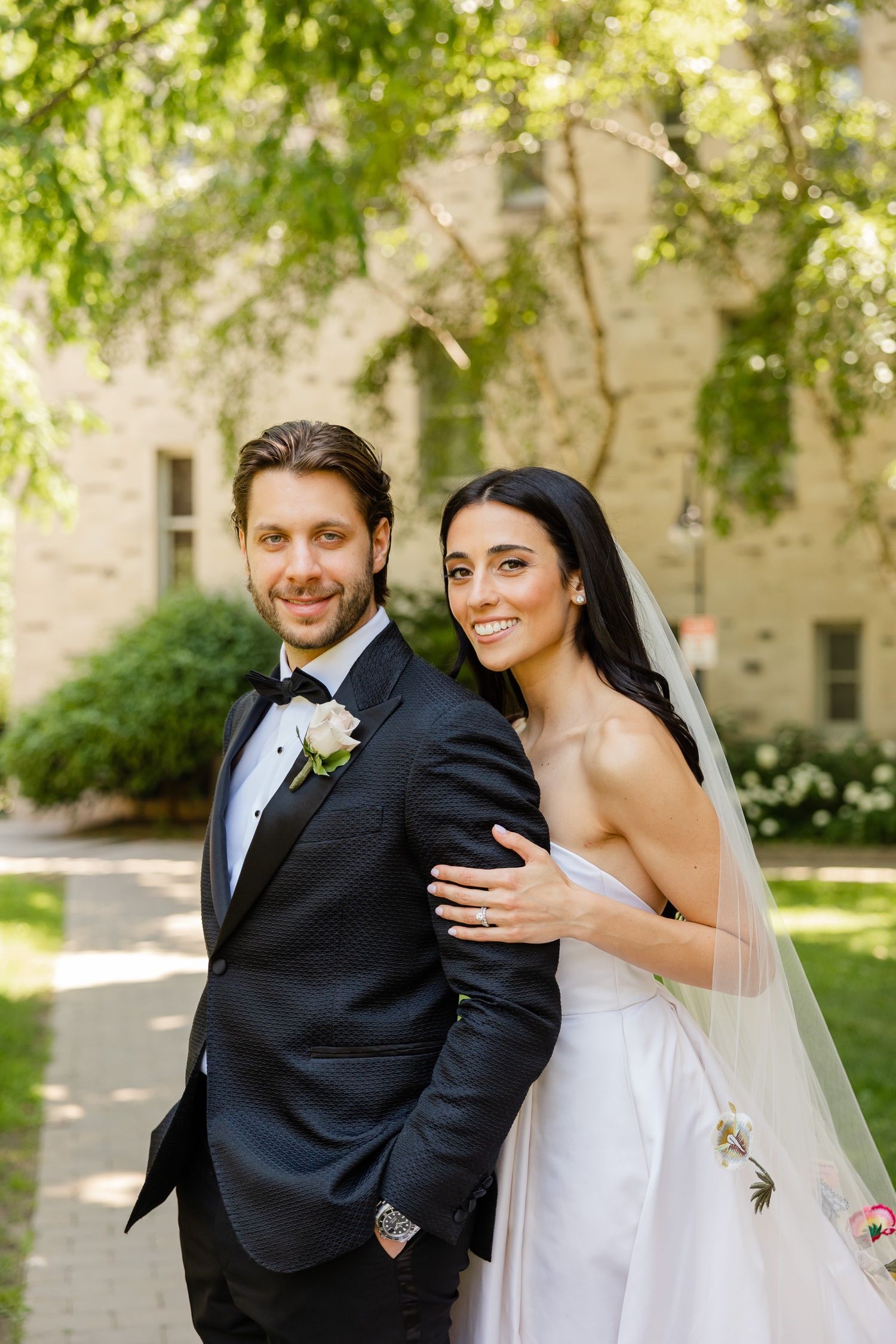 Joyful wedding portrait with couple in formal attire standing against stone building with overhanging trees.