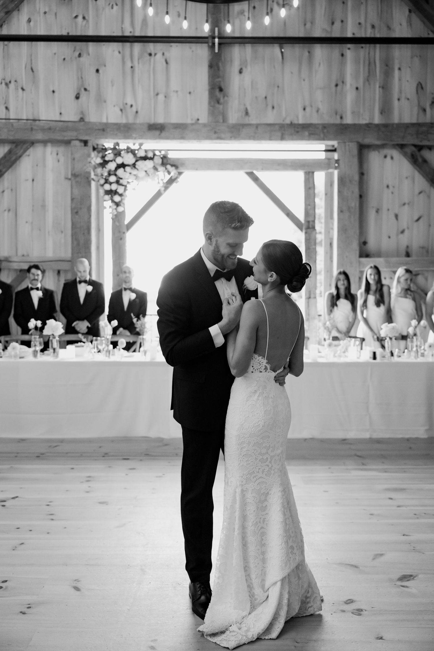 A romantic first dance between newlyweds in a rustic barn venue with string lights and white table settings.