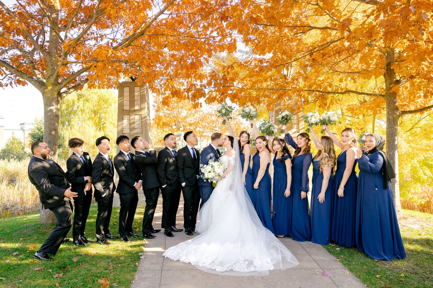 Bridal party poses outdoors with groomsmen in black and bridesmaids in royal blue under fall foliage.