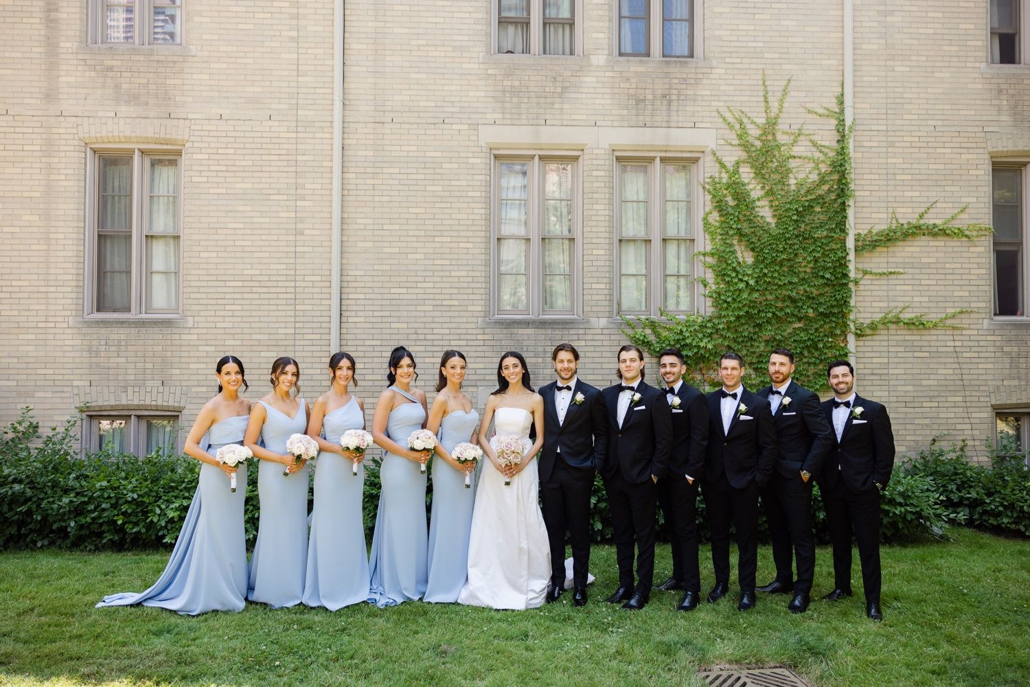 Wedding party poses formally with bridesmaids in blue and groomsmen in black tuxedos.
