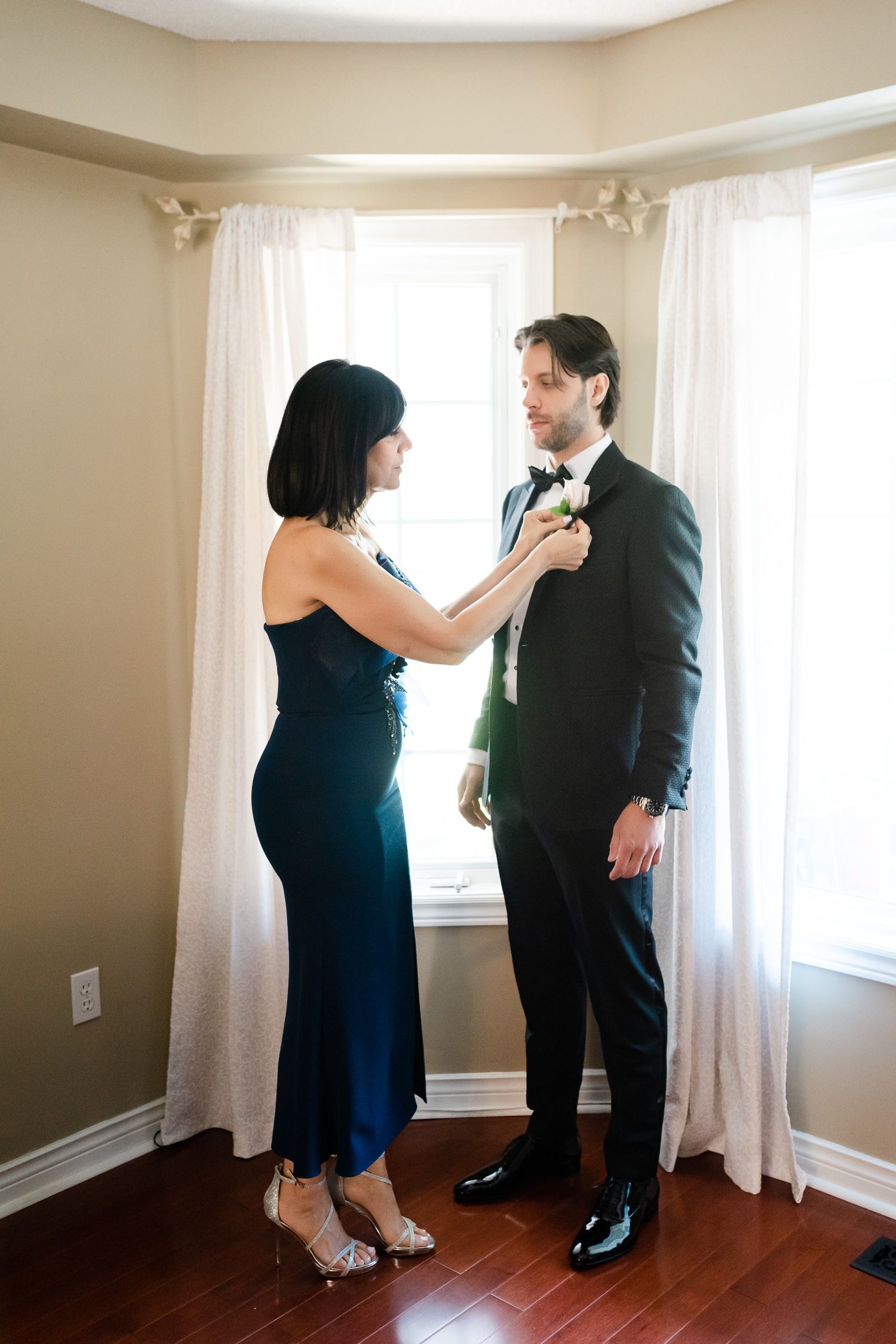 A woman in navy dress helps adjust a bowtie on a man in formal attire near curtained windows.