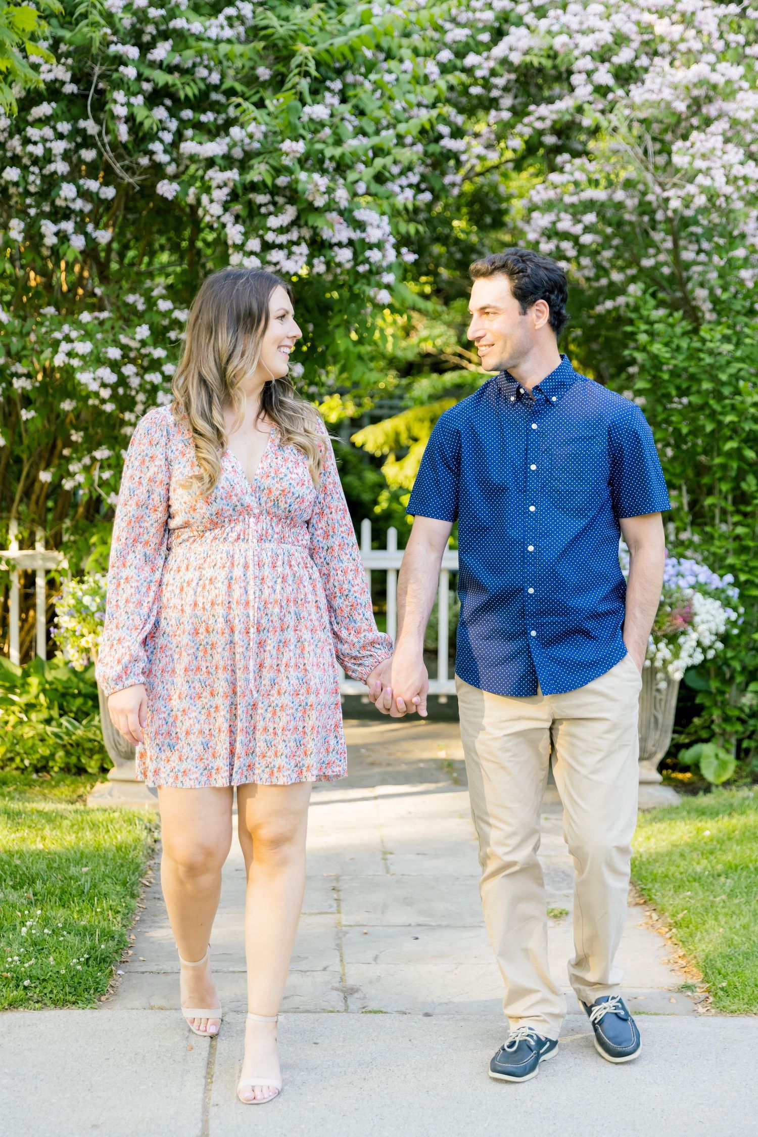 A pair walks together on a garden pathway surrounded by blooming white flowers and lush vegetation.