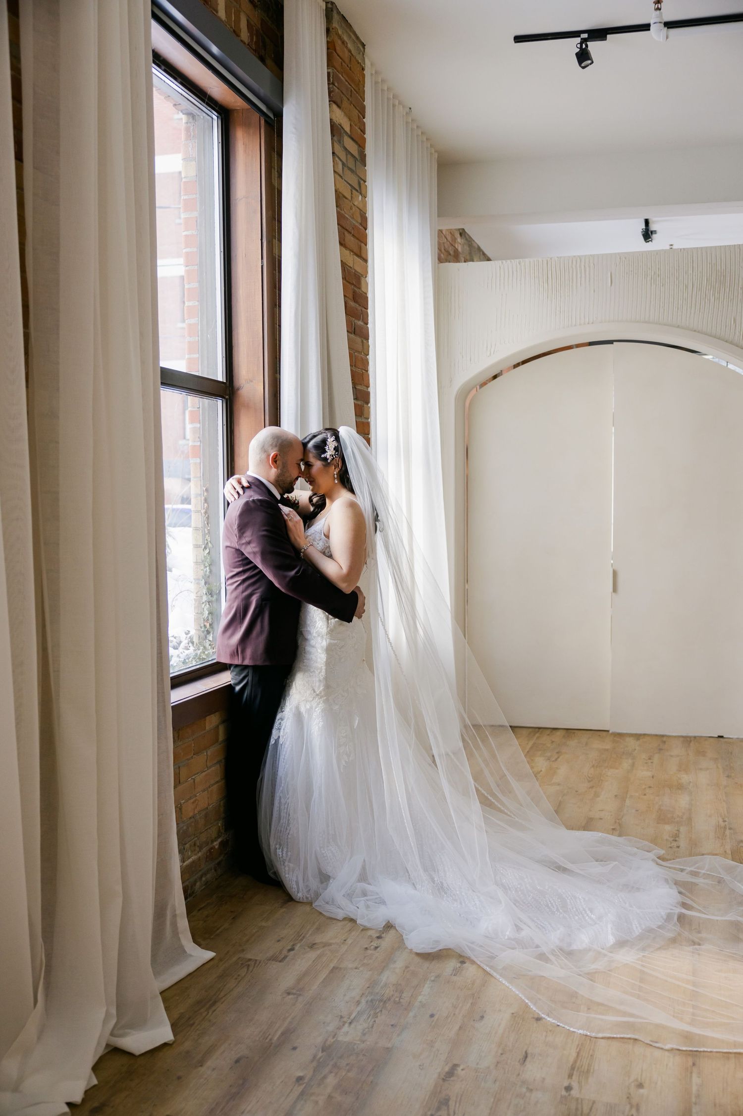 A wedding couple shares an intimate moment by a large window in an elegant indoor space.