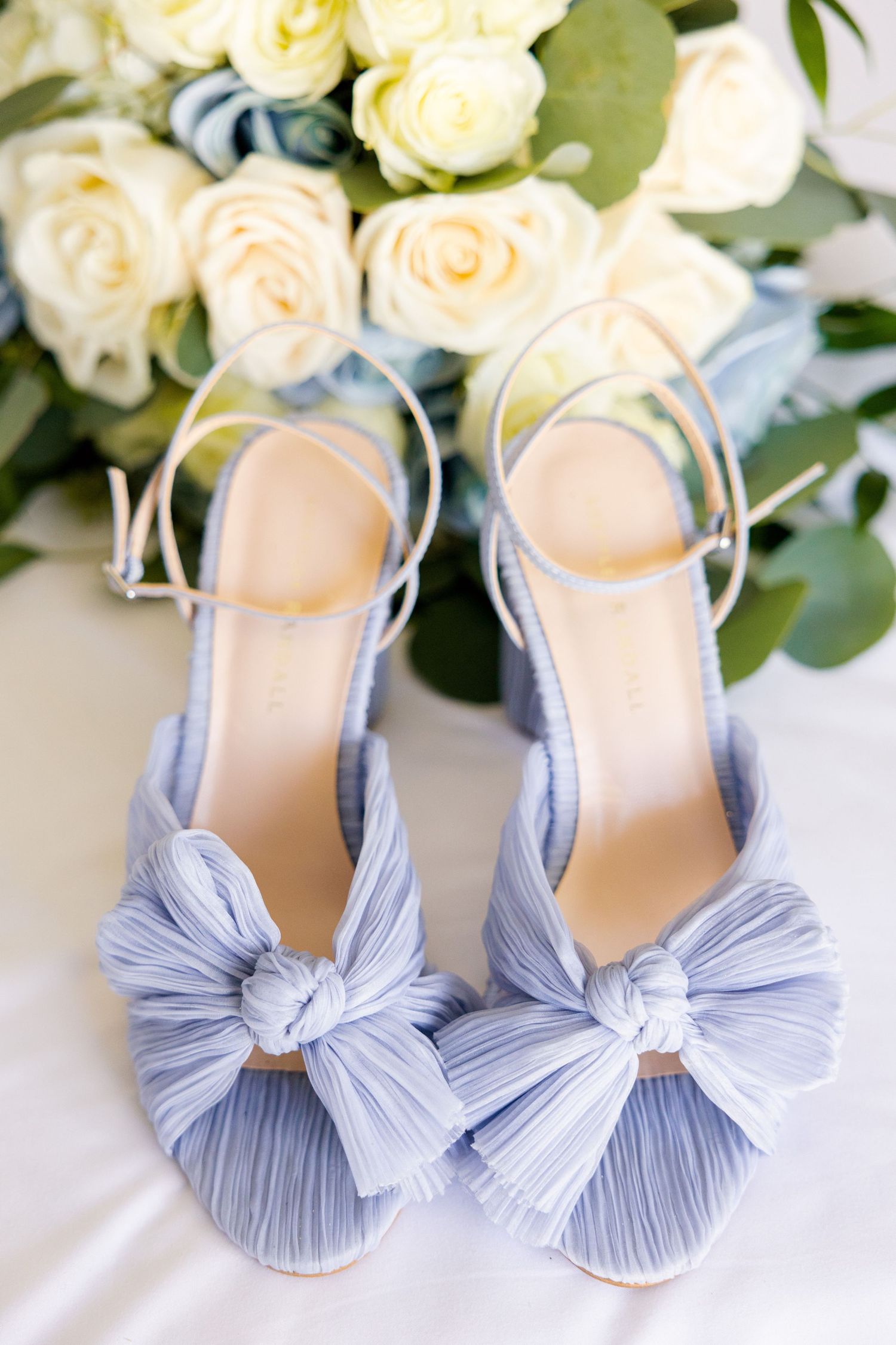 Pale blue pleated bow wedding shoes with ankle straps against white roses and greenery.