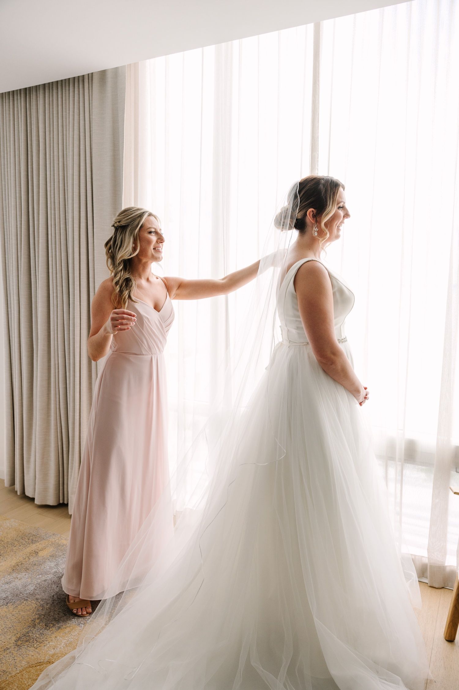 A bridesmaid in a blush gown helps with final adjustments to a white wedding dress by a window.