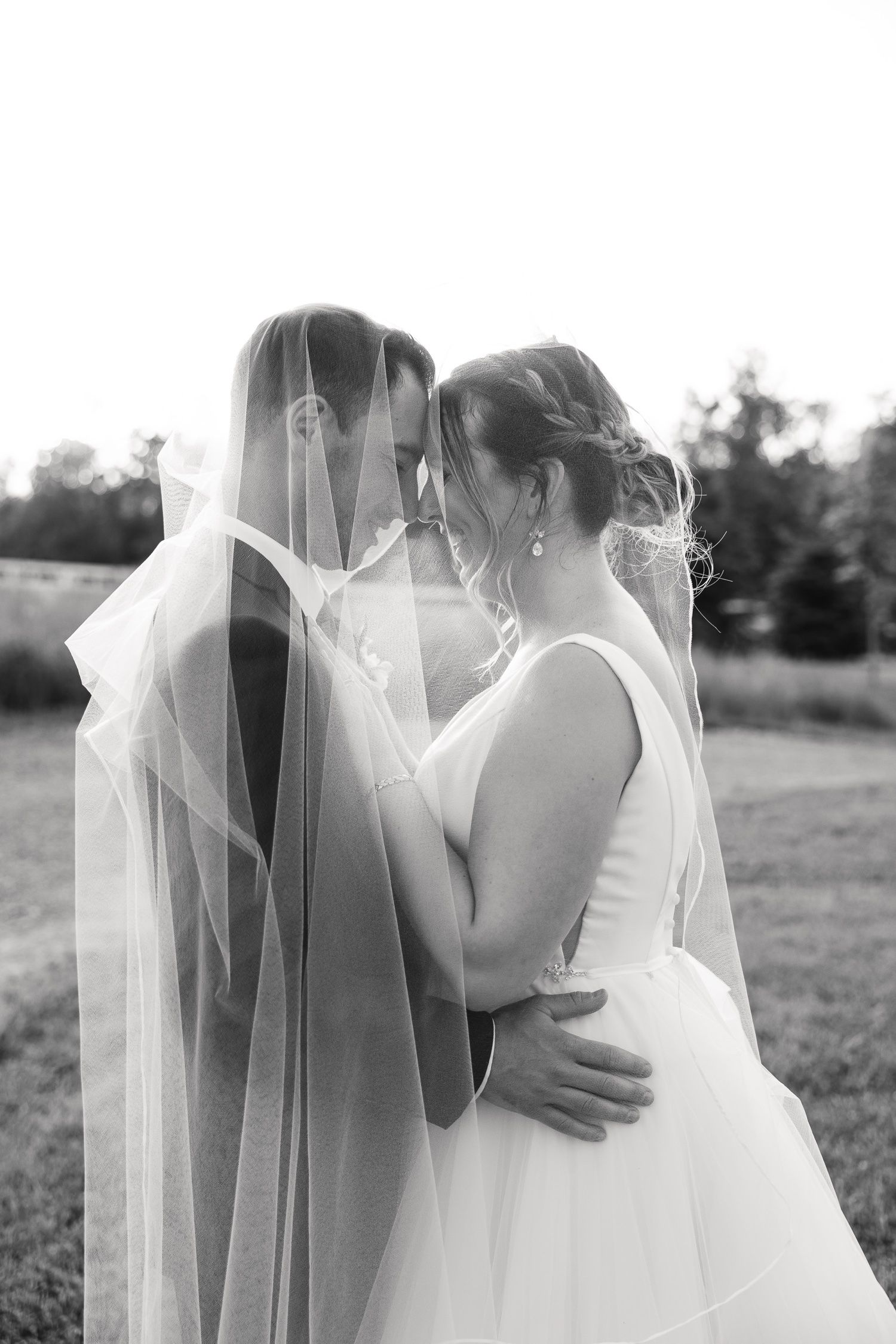 Black and white portrait of couple sharing intimate moment under wedding veil outdoors.