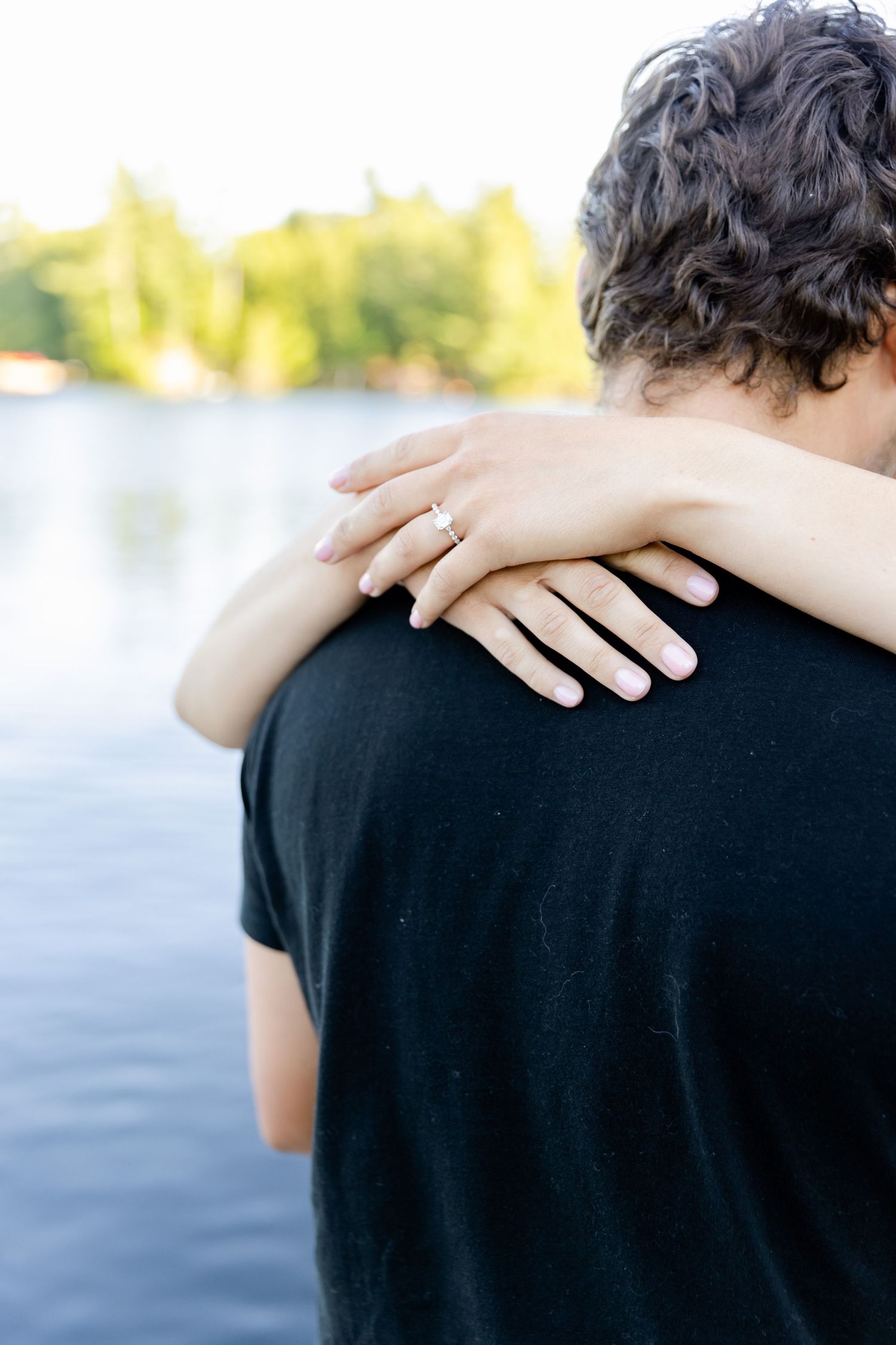 Close-up shot of hands with an engagement ring embracing against a black t-shirt with water in background.