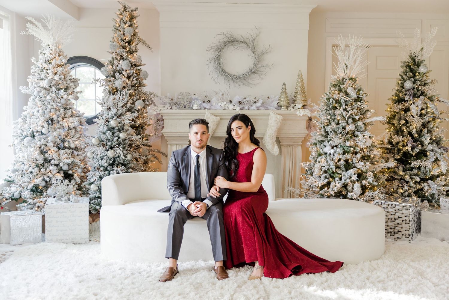 Elegant couple poses in front of snow-flocked Christmas trees and white fireplace mantel in romantic holiday photoshoot.