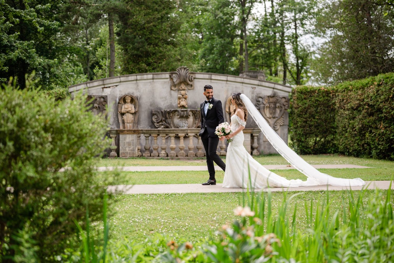 Elegant wedding photo of couple posing by stone wall in landscaped garden setting.
