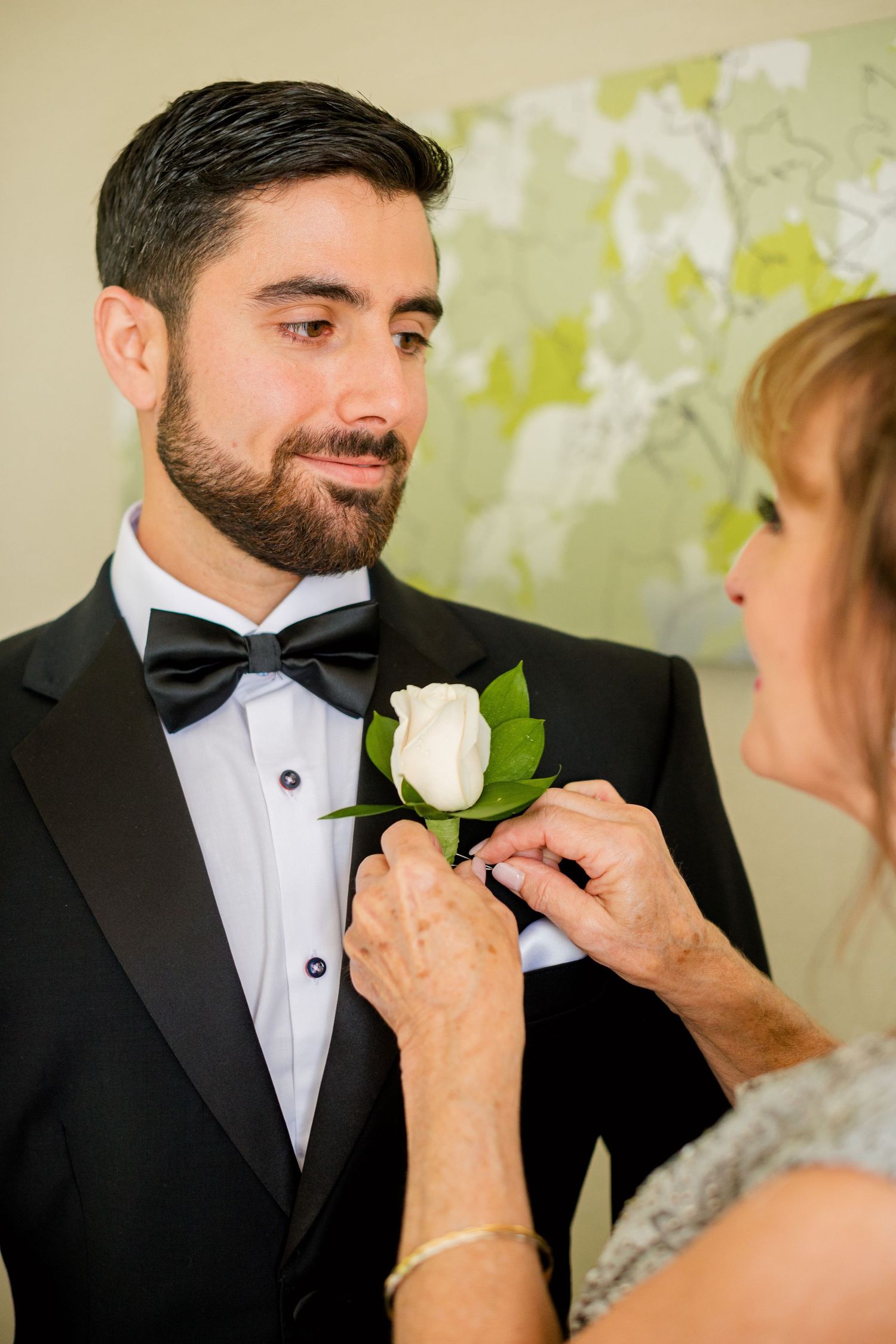 A white rose boutonniere being pinned to a black tuxedo lapel against abstract wall art.