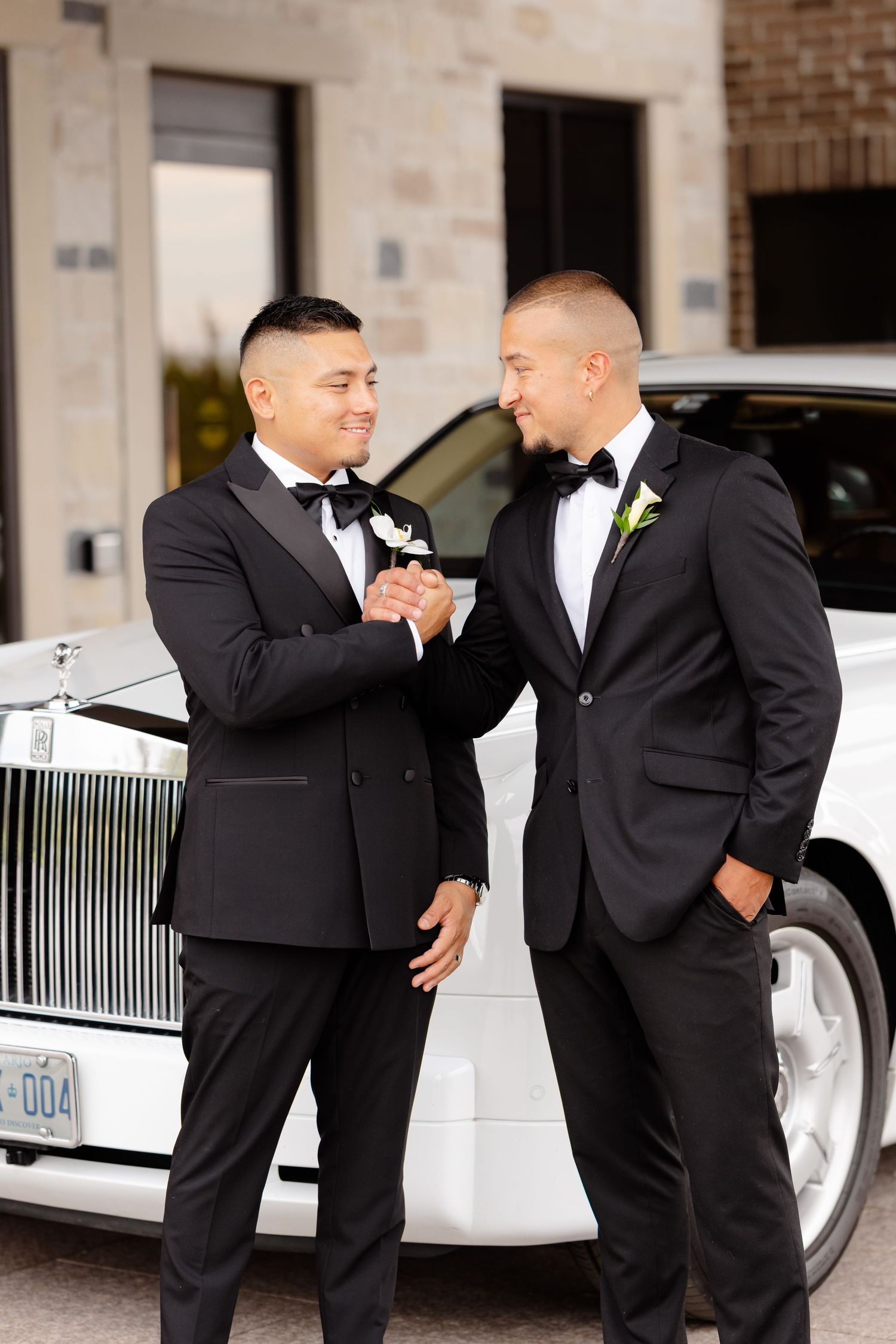 Two groomsmen in black tuxedos and bow ties share a laugh in front of a white Rolls Royce.