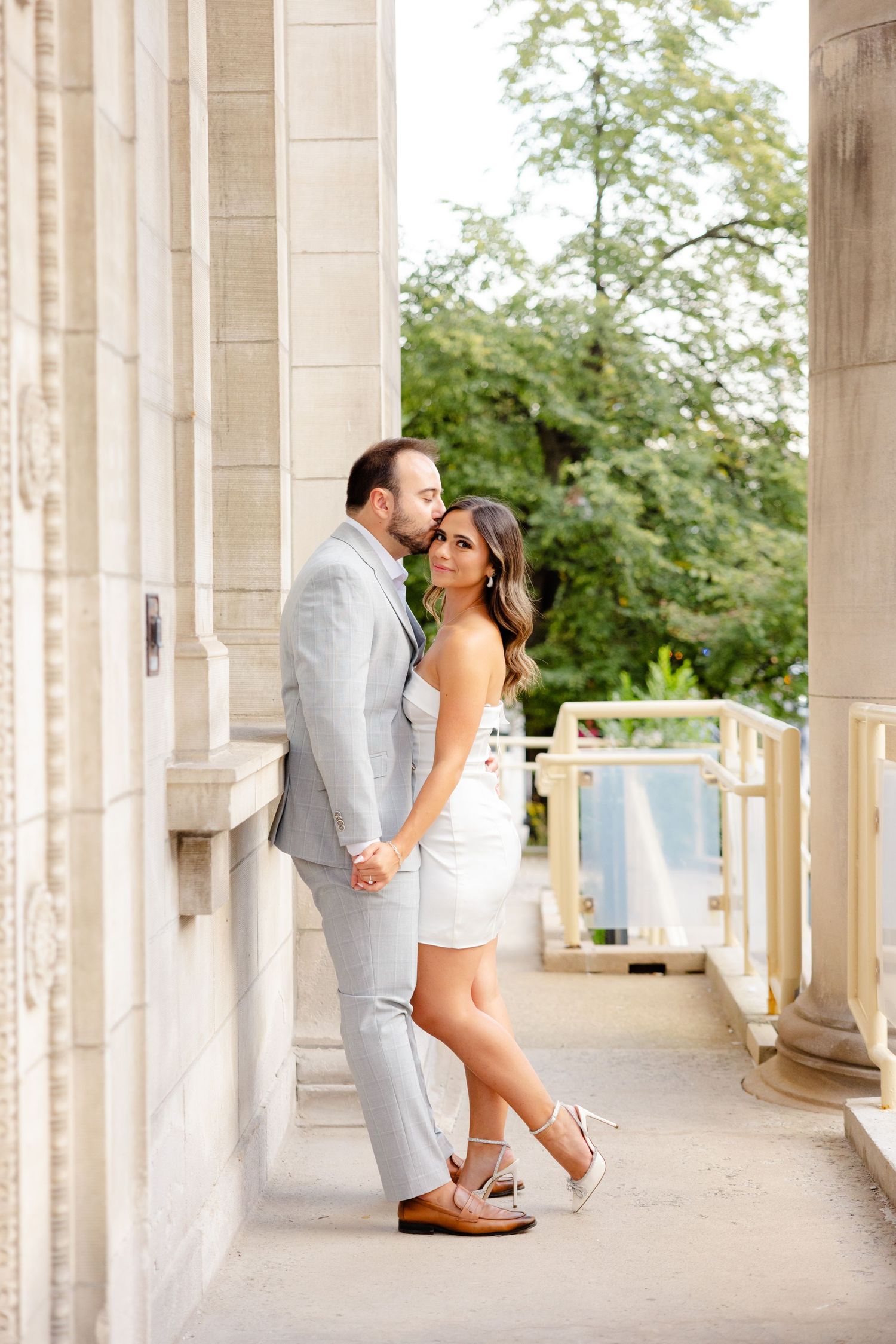 A couple stands close together on a classic stone terrace with columns and trees in view.
