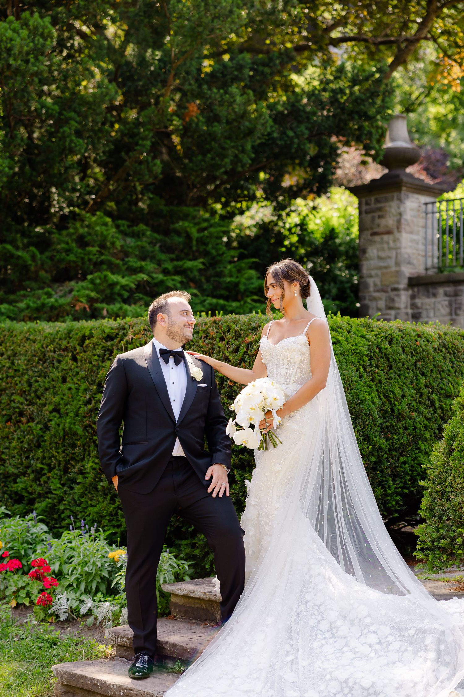A couple in wedding attire poses in a lush garden setting with stone pillars and hedges.