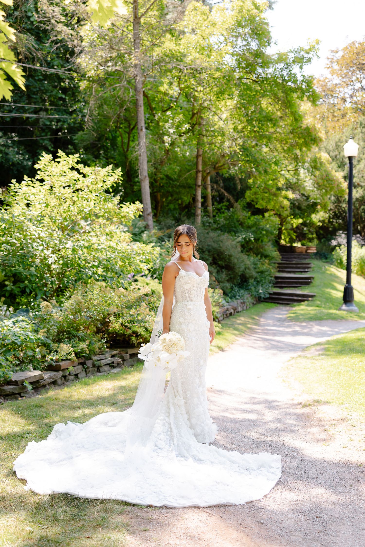 A stunning white lace wedding gown with long train photographed on a garden path surrounded by lush greenery.