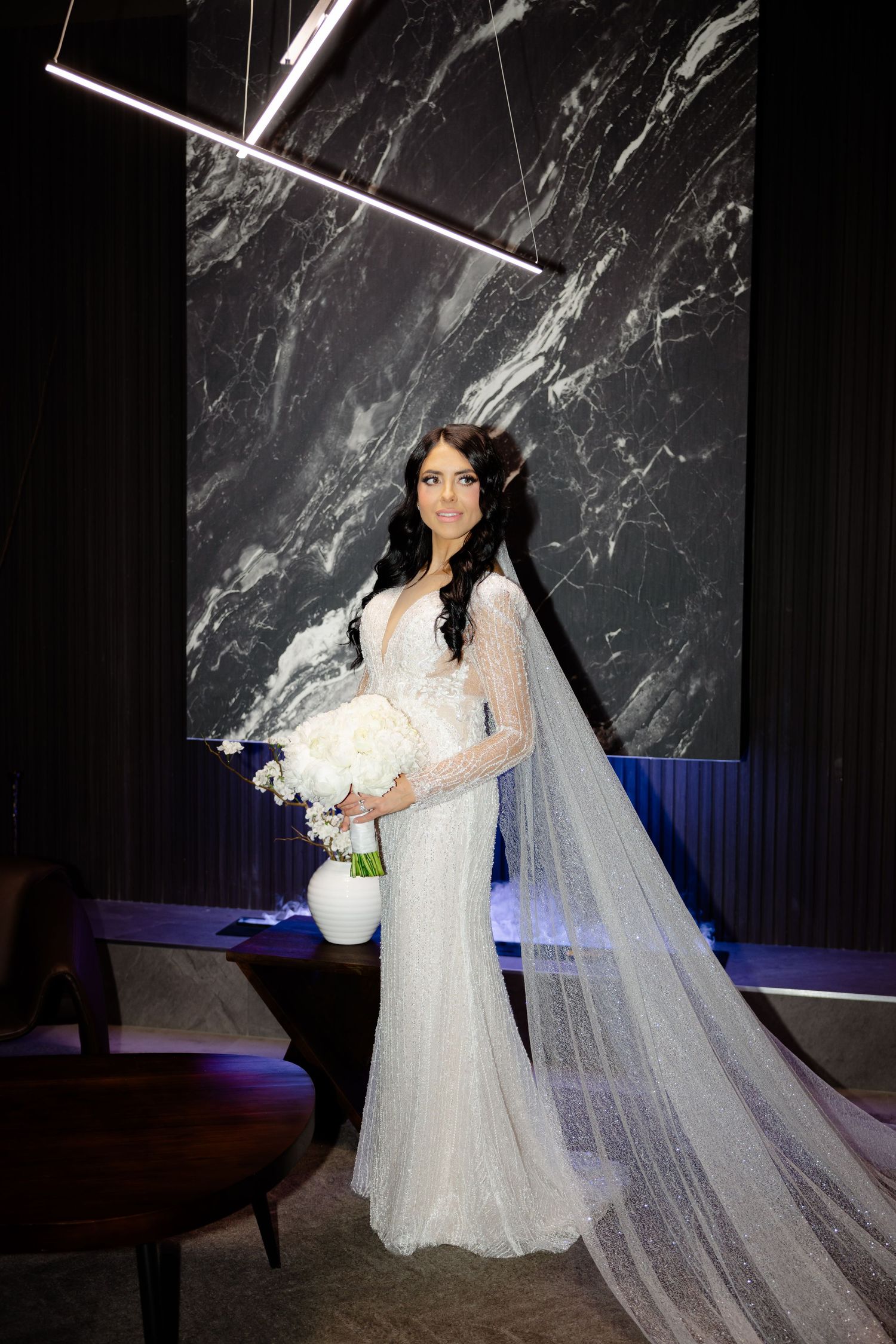 Elegant bride with long veil poses with white bouquet against marble wall with geometric lighting.