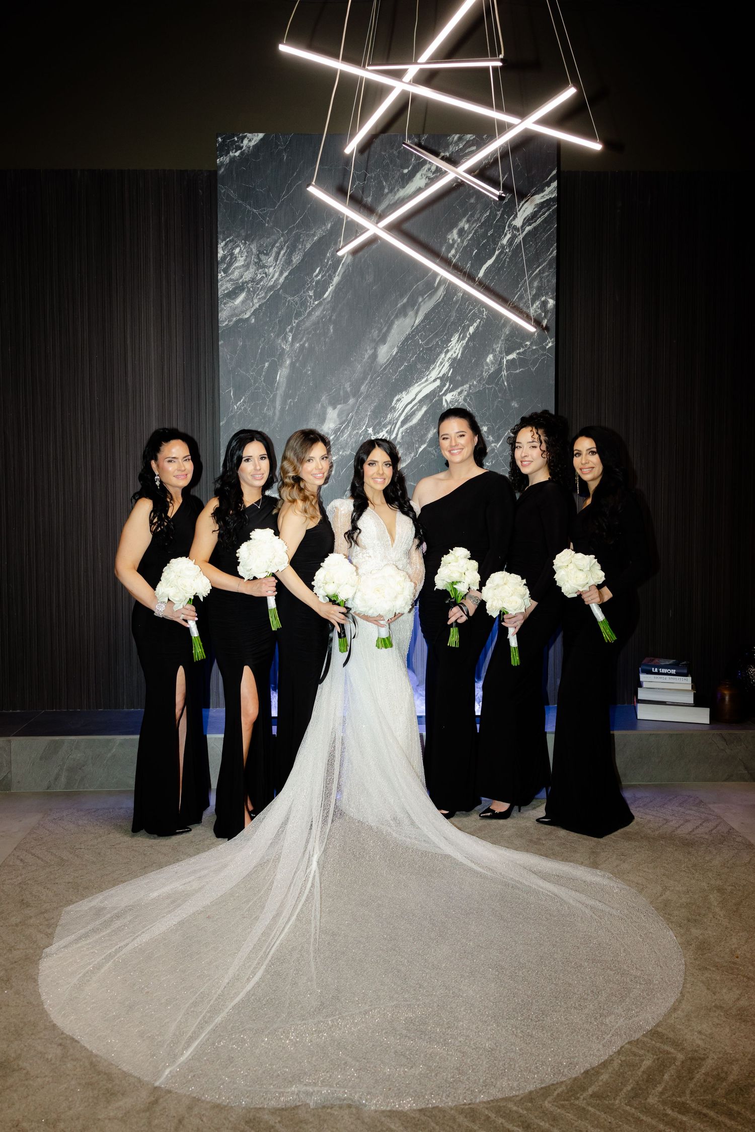 Bridal party in black dresses poses beneath geometric lighting with bride's flowing train.