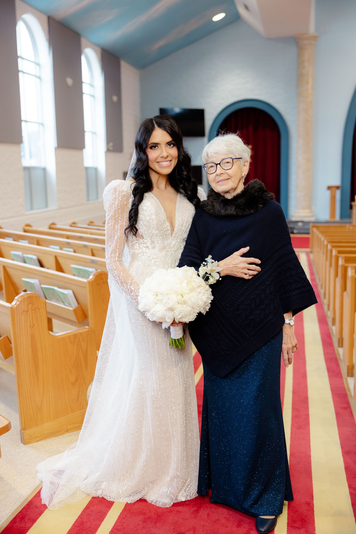 Elegant bride in lace gown poses with elderly guest wearing navy dress inside traditional church sanctuary.
