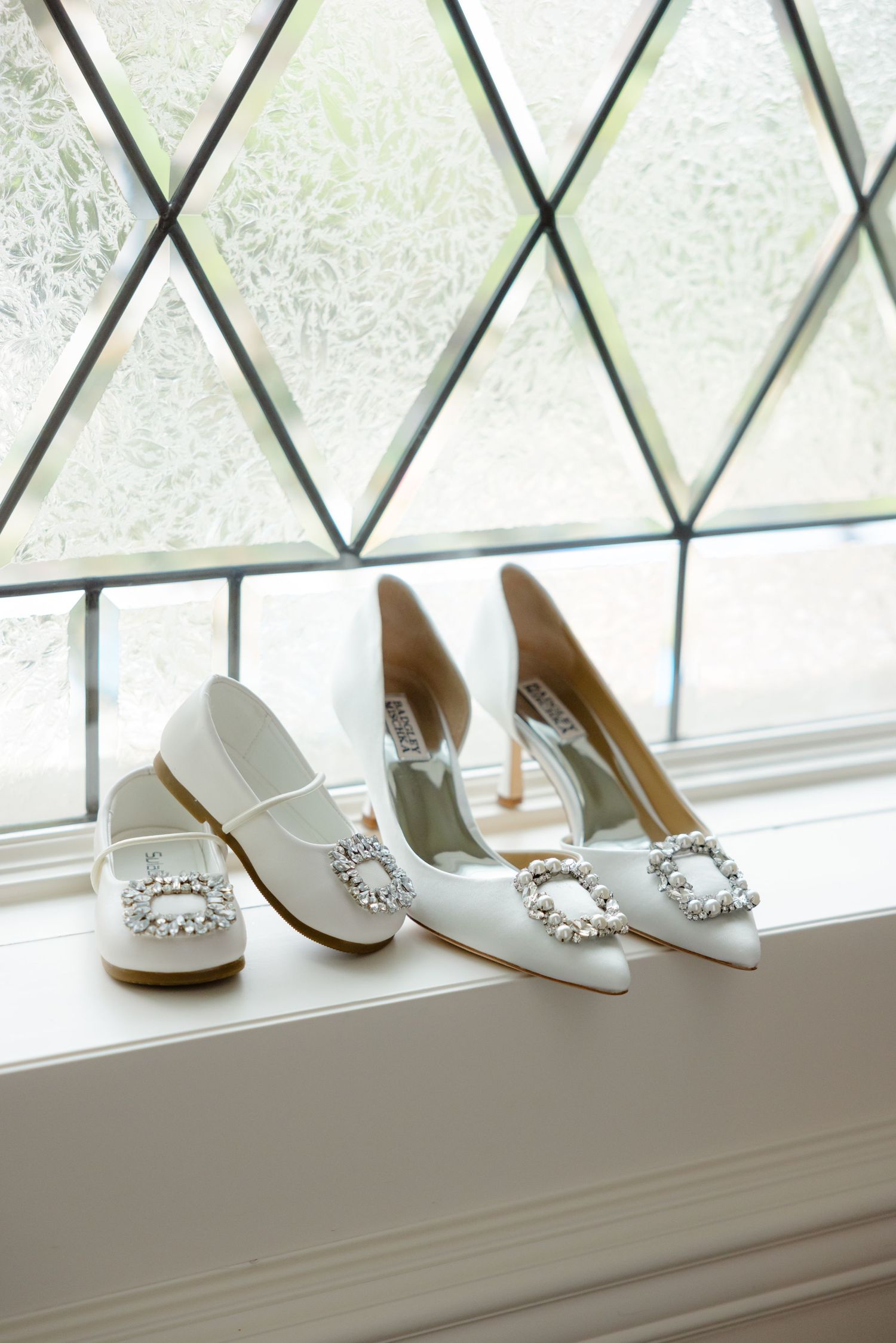 Two pairs of white bridal shoes with crystal buckles displayed on windowsill.