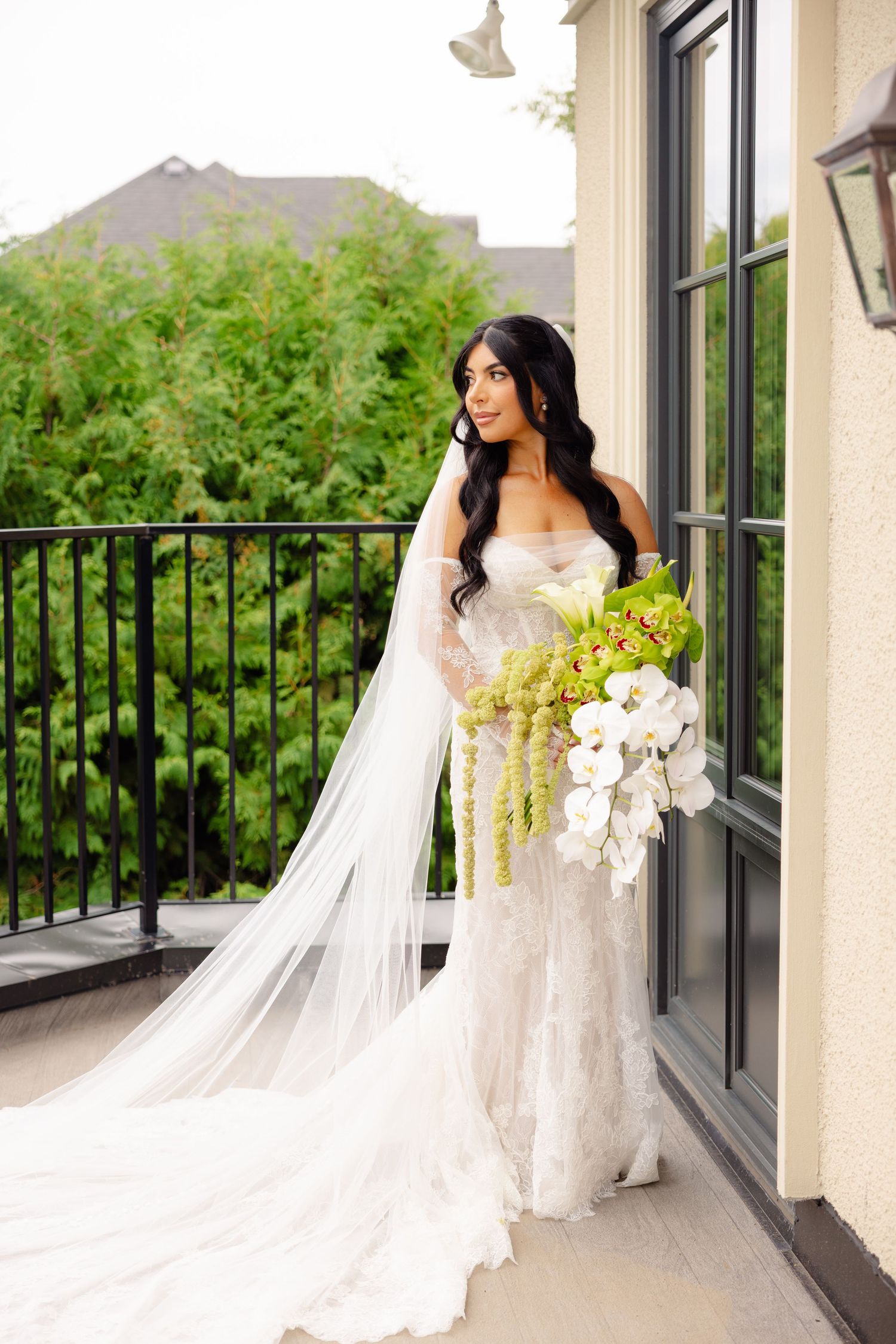 A bride in a flowing white lace gown and cathedral veil poses on an elegant balcony with mountain views.