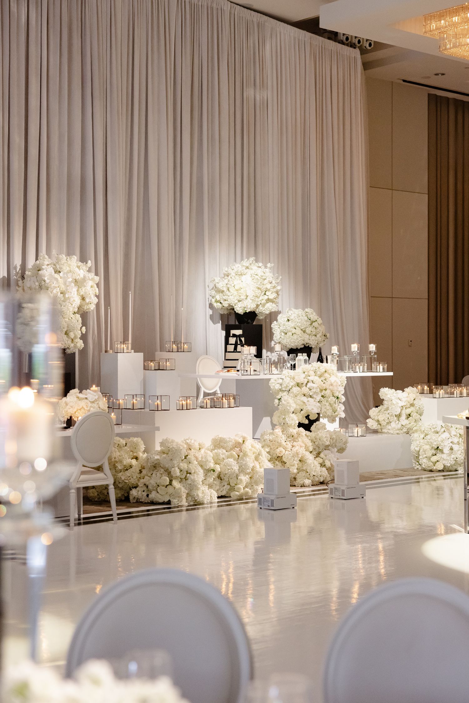 White floral centerpieces and candles line a modern wedding reception head table with elegant draping and white chair decor.