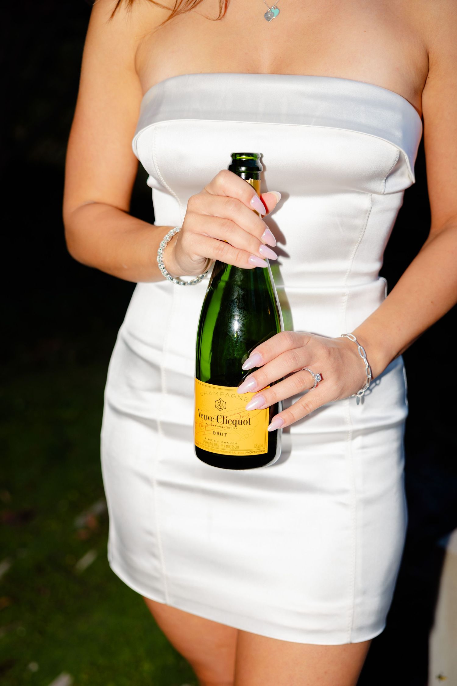 Person in white strapless dress holding a bottle of Veuve Clicquot champagne against dark background.