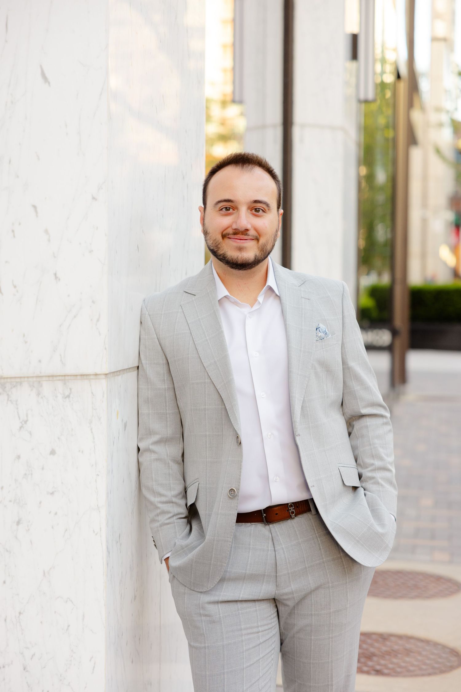 Professional portrait of a man in a light gray suit and white shirt standing against a white column.