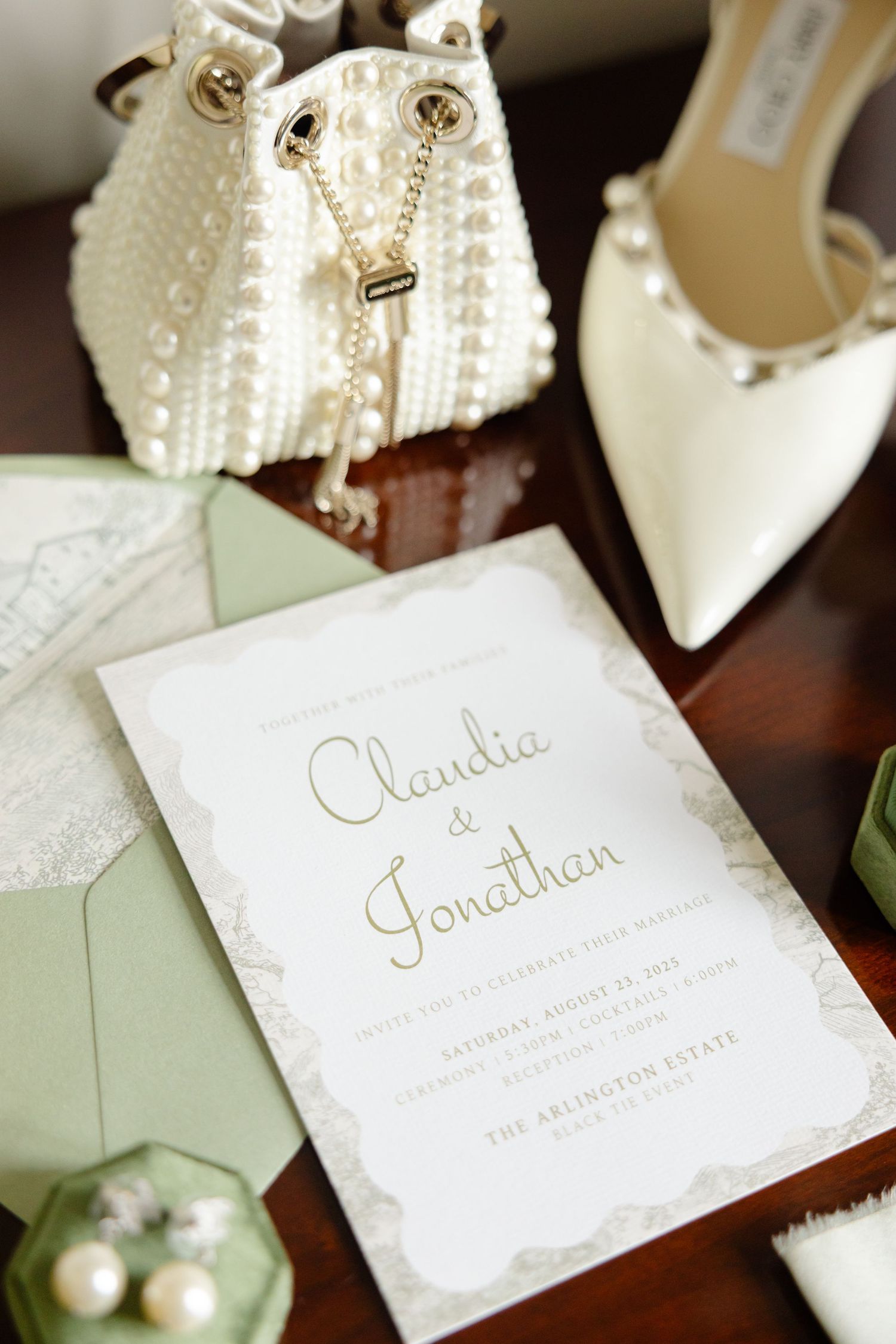Elegant white wedding invitation with sage green envelope displayed alongside pearled handbag and bridal heels.