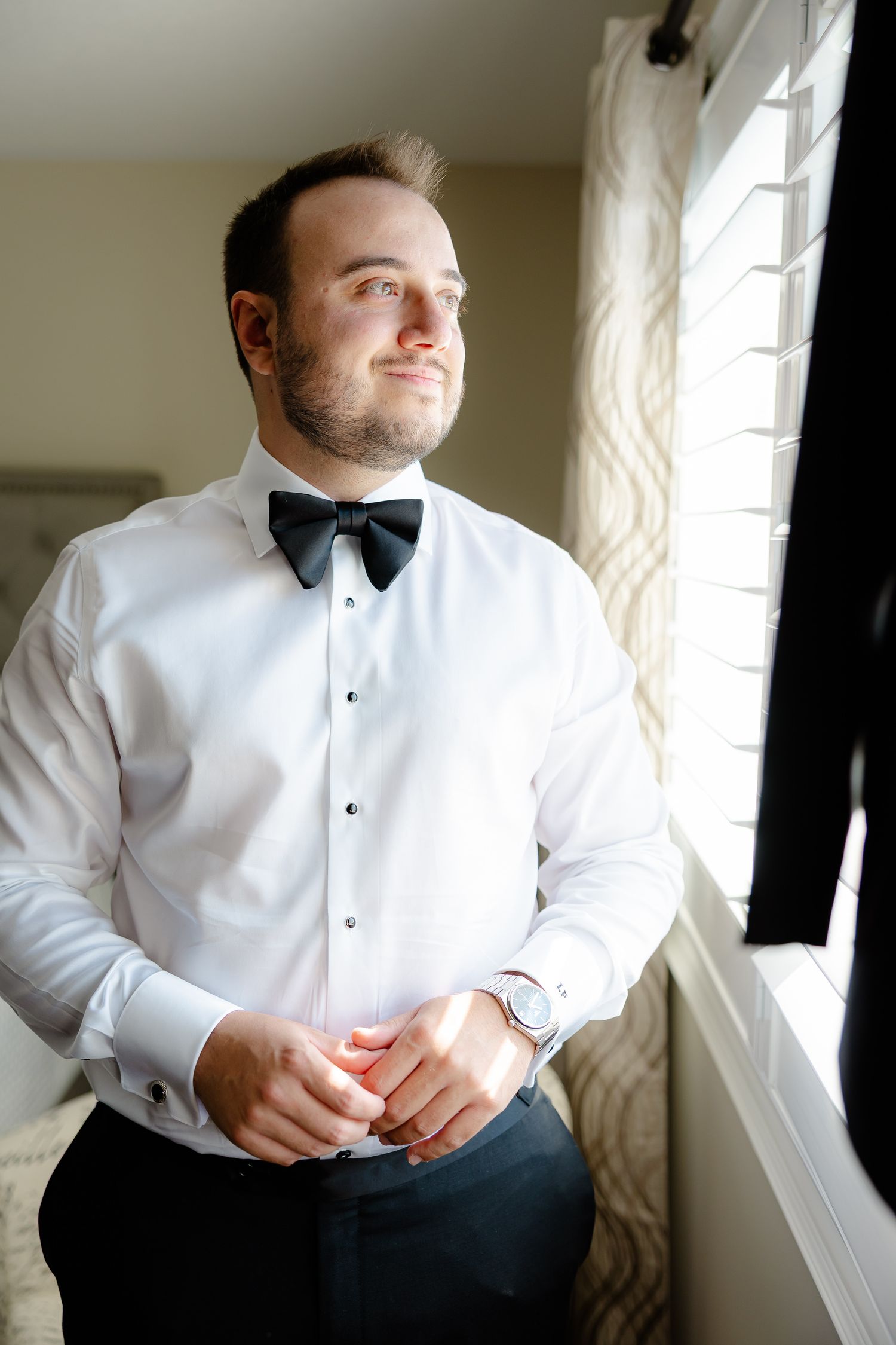 A groom in a white dress shirt and black bow tie stands by a window while getting ready for a formal event.