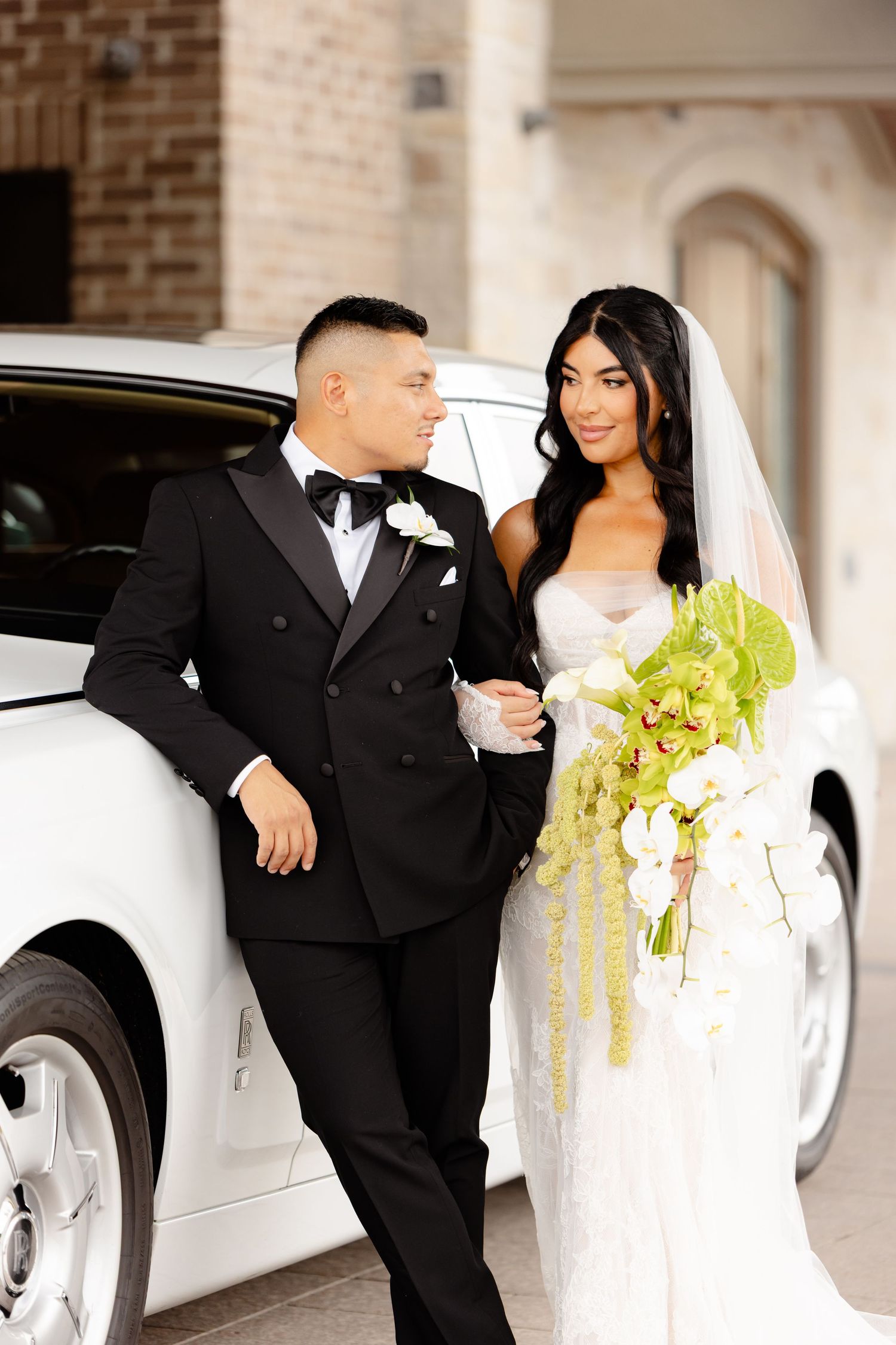 Newlywed couple poses elegantly next to white luxury car, with bride holding cascading green and white bouquet.