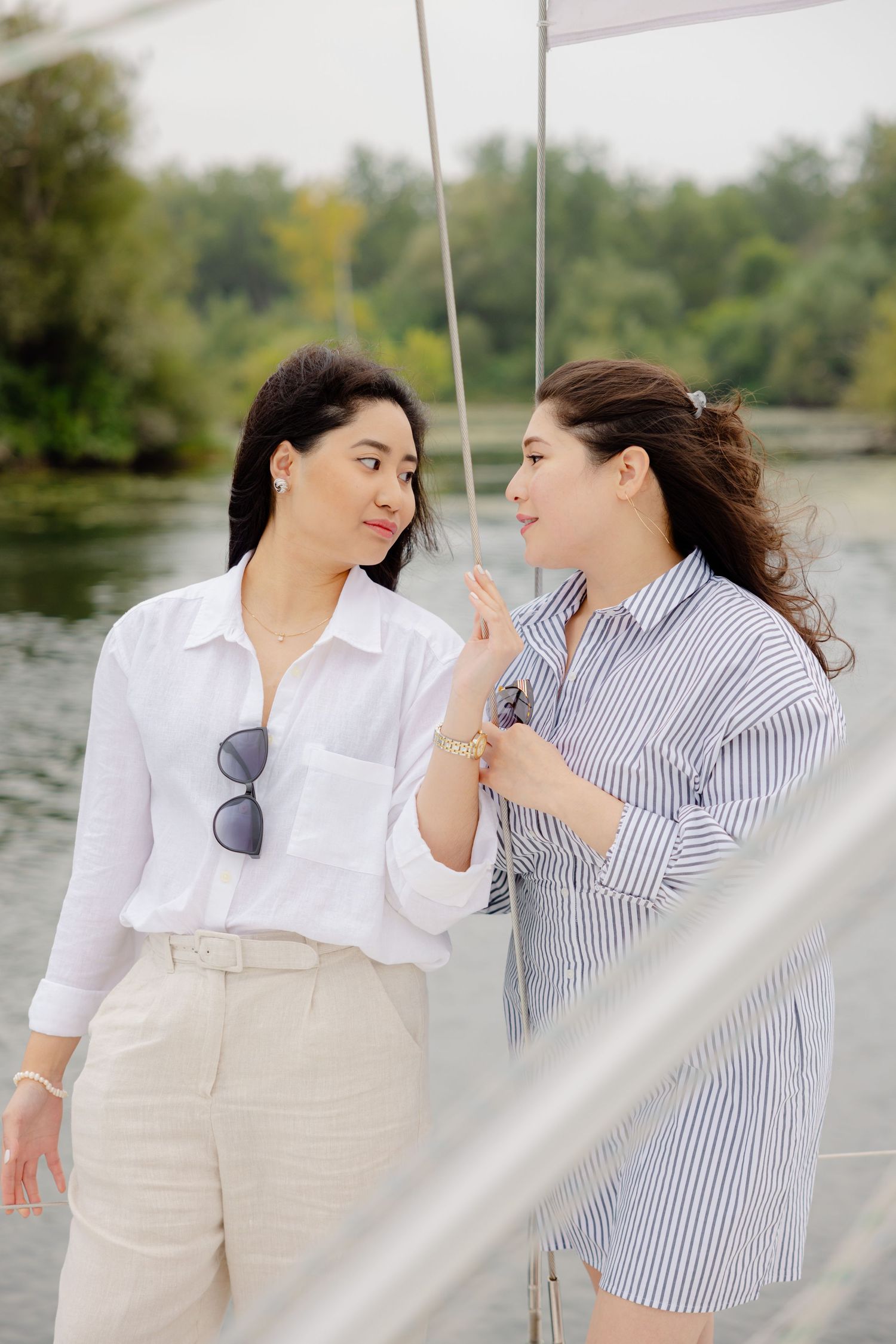 Two women in casual attire stand close together on a boat dock with a serene lake and trees in the background.