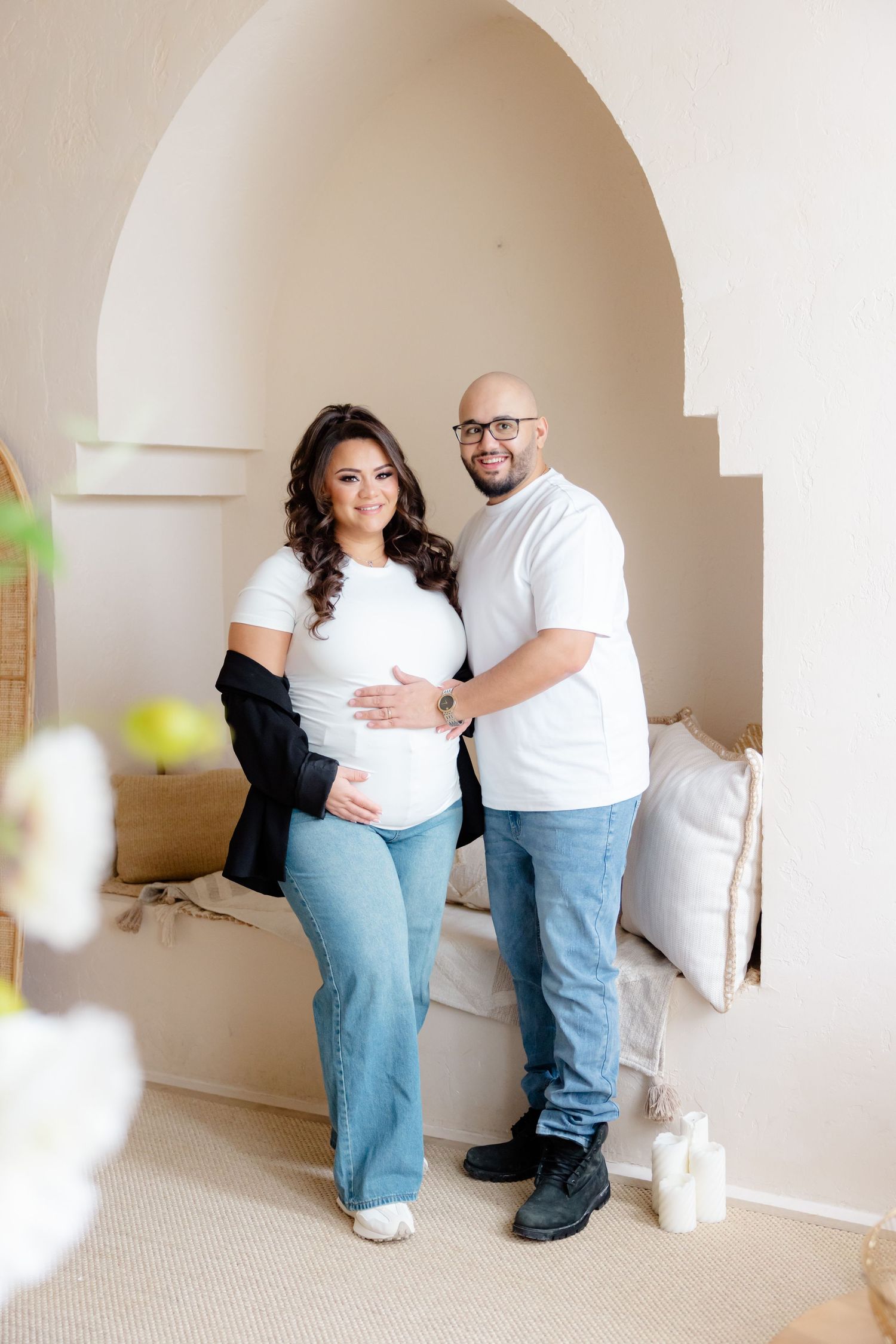 A couple in matching white t-shirts and jeans embrace under an arched doorway in a light, neutral-toned room.