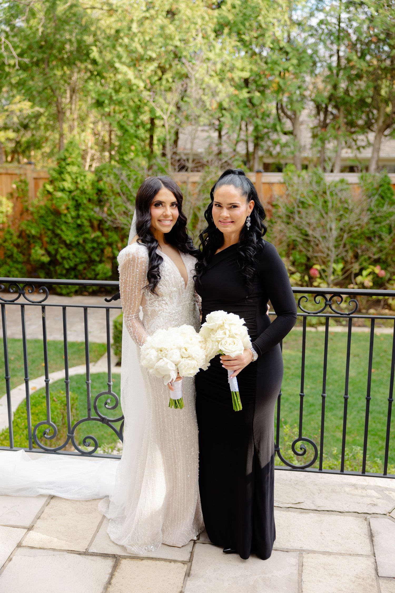 Two women in contrasting white and black formal dresses hold white bouquets on outdoor stone patio.